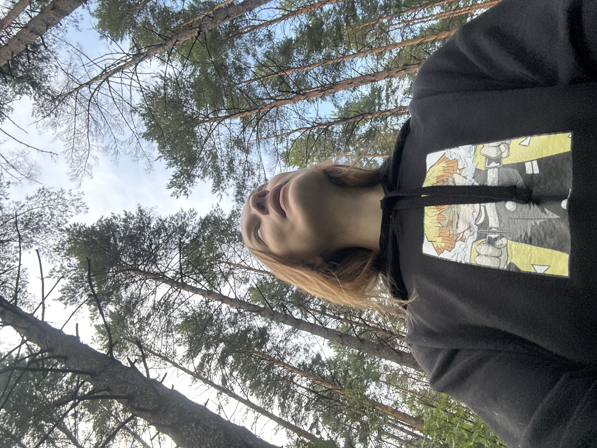 The image is a low-angle, outdoor selfie taken in a forest with tall pine trees. The subject is a person with shoulder-length blonde hair, wearing a black hoodie with a graphic print of an anime character on the front. The person is looking upwards, and the sky is visible through the tree canopy, with patches of blue sky and clouds. The forest floor and surrounding vegetation are not clearly visible due to the low-angle shot and the focus on the trees and the subject. The location is specified as Chaikovsky, Russia.