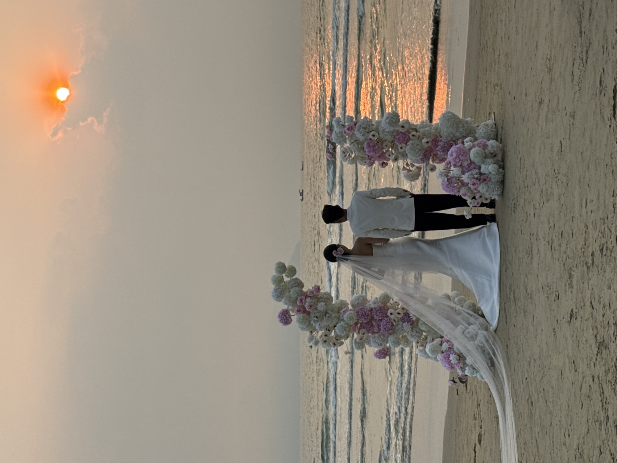 A wedding ceremony on a beach at sunset. A bride in a white dress and veil stands next to a groom in a white shirt and dark pants. They are positioned in front of a floral arch of white and pink flowers. The sun is setting in the sky, visible on the left, casting a warm glow on the water and sand. The location is likely a beach in Đà Nẵng, Vietnam.