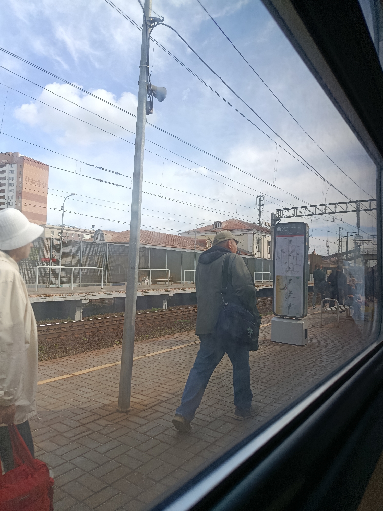 The image shows a train station platform in Pavlovsky Posad, Russia. A man wearing a green jacket and a cap is walking past a public information board that displays a schedule. He has a dark bag slung over his shoulder. To the left, a person in a white hat and jacket is standing, holding a red bag. The platform is paved with brick, and train tracks are visible in the background. Overhead, electrical wires for the train are strung across the sky, which is partly cloudy. A tall building and other structures are visible in the distance. The photograph is taken from inside a train looking out through the window.