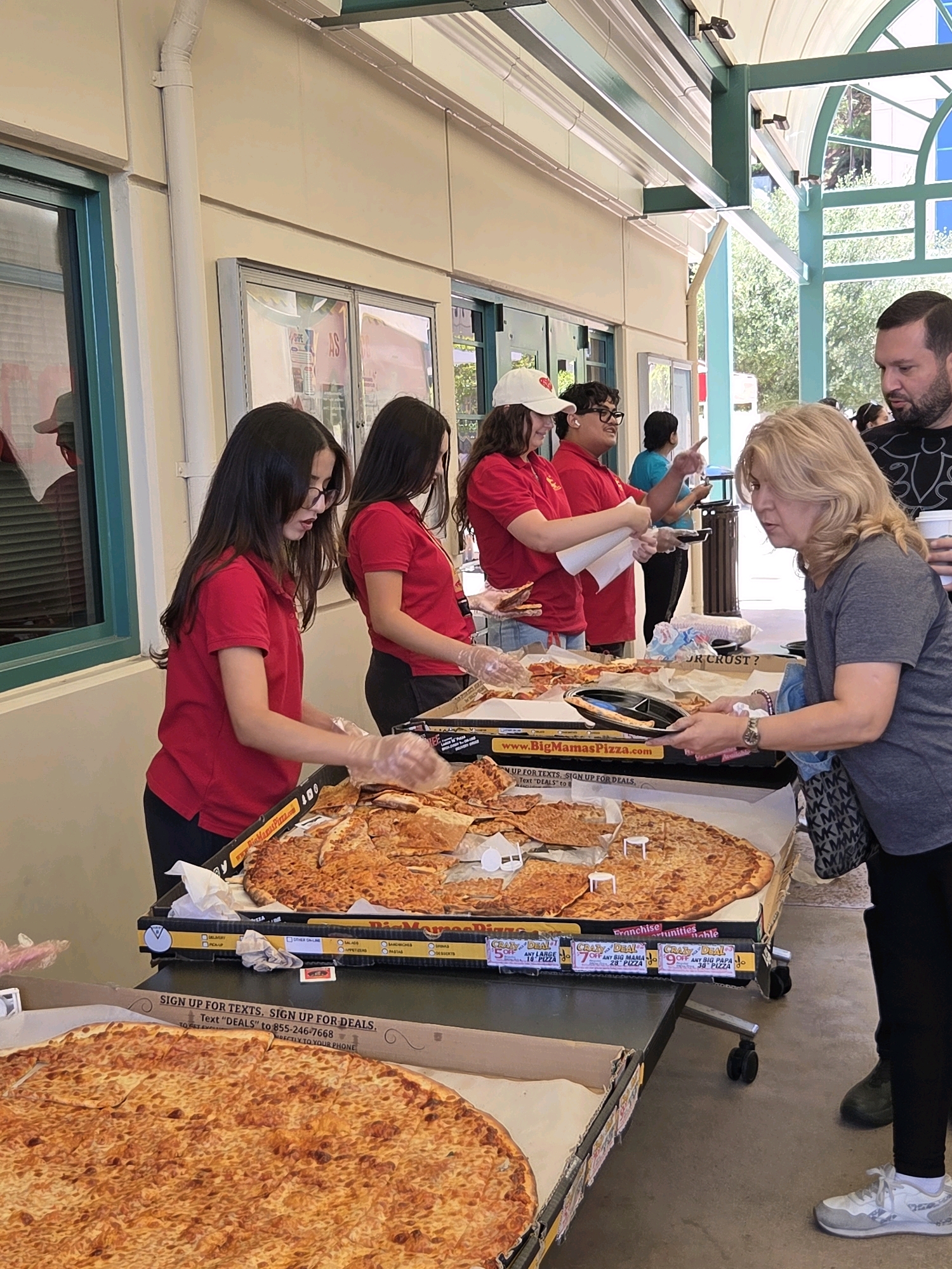 In Glendale, California, a scene unfolds outdoors at what appears to be a pizza sale, possibly a fundraising event, under a shaded awning. Several people, mostly young women in red polo shirts, are serving slices of cheese pizza from large boxes arranged on a table. One young woman in a red shirt and glasses, wearing clear plastic gloves, is holding a slice of pizza, looking towards a blonde woman in a gray t-shirt who is reaching out with a black plate. Another young woman in a red shirt and a white baseball cap stands beside the first, also wearing gloves and holding a piece of pizza. Behind them, a young man in a red shirt and glasses gestures with his right hand while talking. To the right, a man with dark hair and a beard, wearing a black t-shirt with a white design, holds a white cup and observes the transaction. The background shows architectural elements of a building with windows, and beyond that, trees and greenery. The pizzas are displayed in cardboard boxes, some with promotional signs. The overall atmosphere suggests a public event focused on selling pizza.