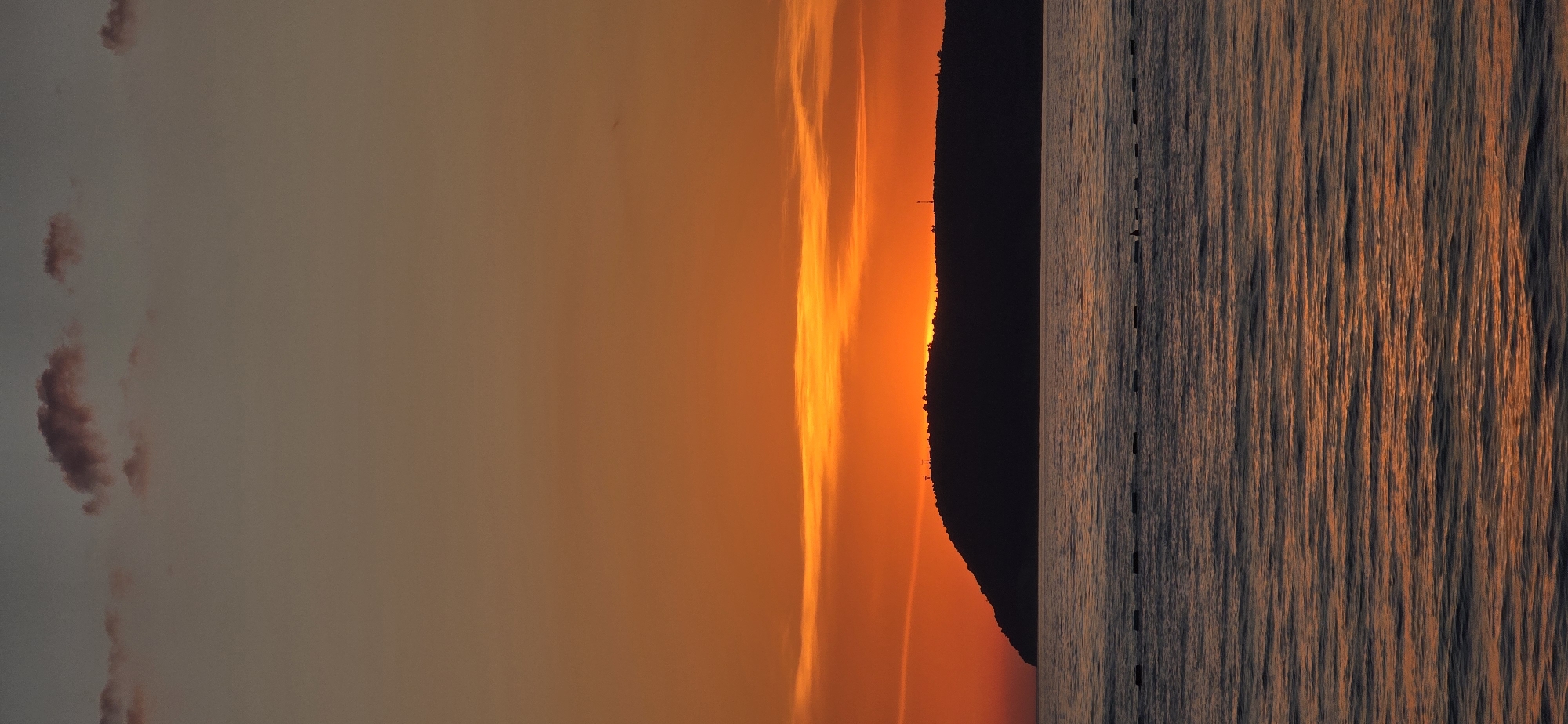 The image depicts a sunset in Bar, Montenegro, with the sun setting behind a dark, silhouetted mountain or landmass. The sky is filled with hues of orange and yellow. To the right is the sea, with the water reflecting the orange light of the sunset, creating vertical patterns of light and dark. There are dark, vertical structures that seem to have been lined up on the surface of the water. There are also a few small, indistinct dark figures near the mountain silhouette.