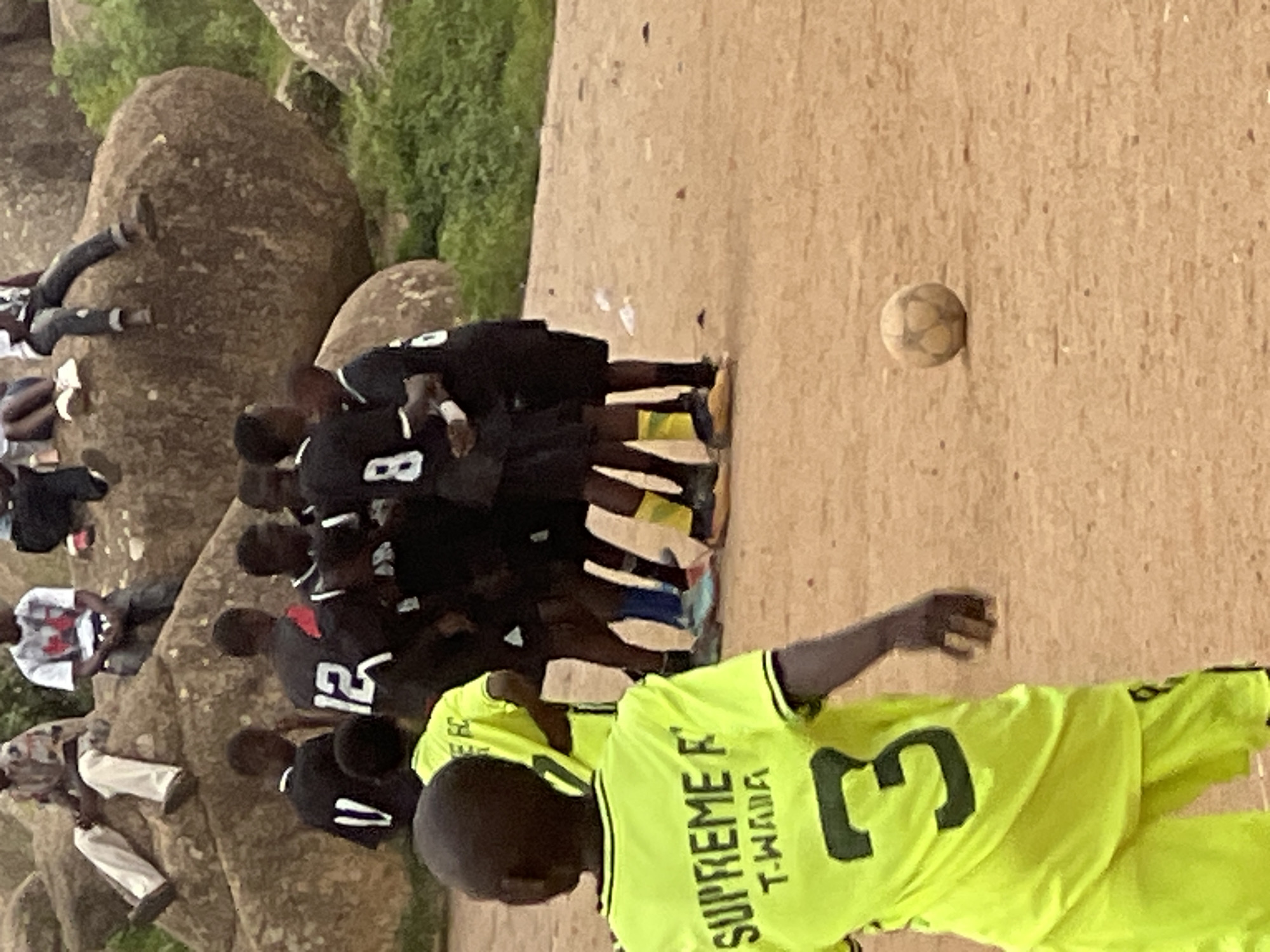 The image depicts an outdoor scene in Jos, Nigeria, likely on a football pitch. In the foreground, a young boy in a bright yellow jersey with "SUPREME FC T-WADA" and the number "3" on the back is seen from behind, facing away from the camera. He is wearing matching yellow shorts. A soccer ball rests on the dirt ground to his right. In the mid-ground, a group of boys in black jerseys and shorts, some with numbers visible like "12" and "8", are gathered. They appear to be participating in a football match. In the background, several individuals are seated and standing on large, rocky outcrops, some appearing to be spectators. The environment is characterized by the dirt ground and sparse vegetation visible behind the rocky formations. The lighting suggests it is daytime.