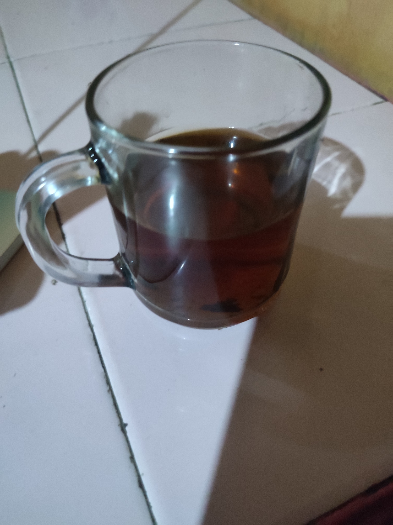 A glass mug filled with dark liquid, possibly tea or coffee, sits on a tiled surface. The mug has a handle and is partially illuminated by a light source creating shadows. The background appears to be a wall. The scene is likely indoors in a home or cafe environment. The image does not provide any specific information that can be used to determine its location in Nusawungu, Indonesia.