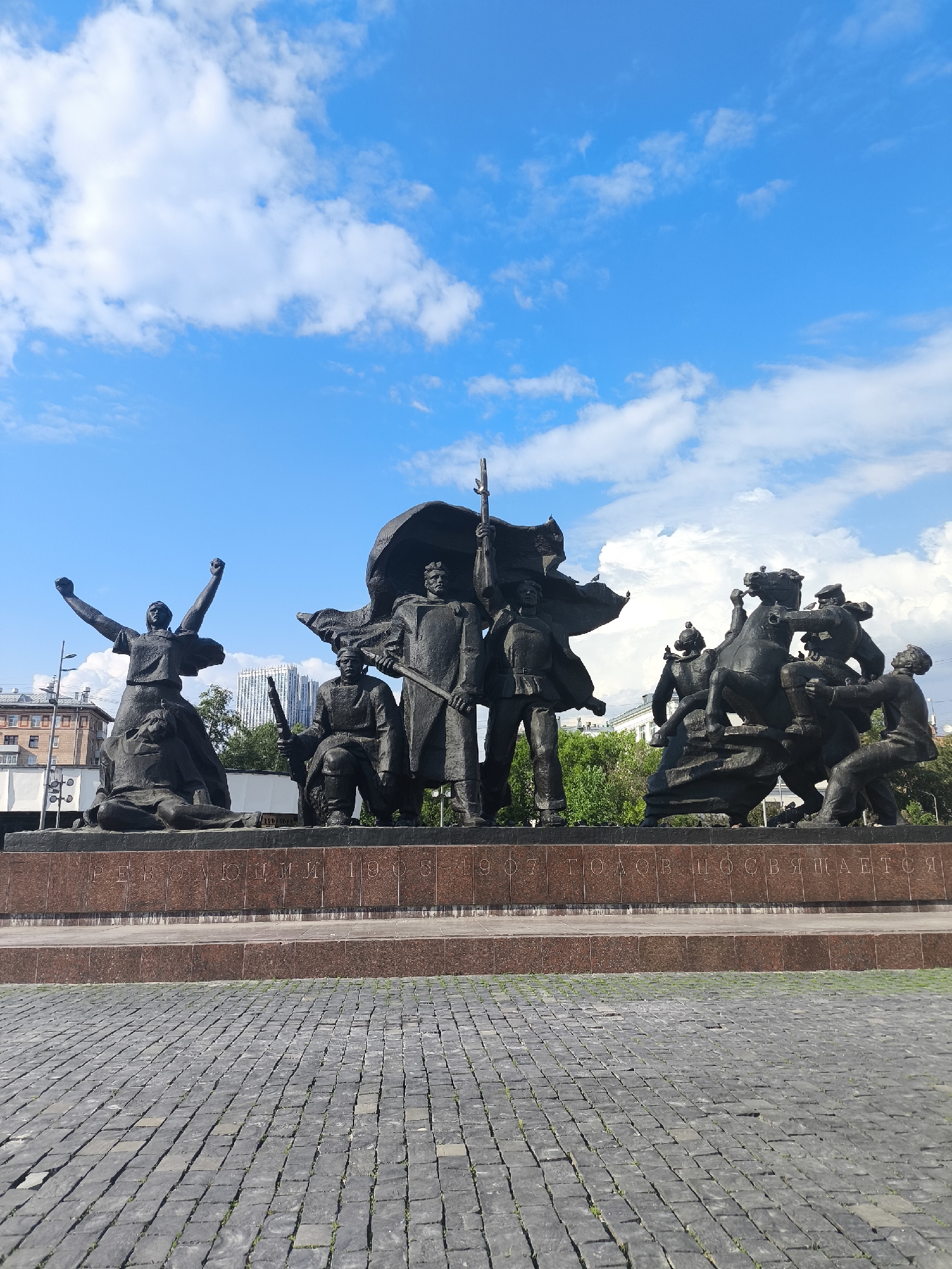The image depicts the Monument to the Revolution of 1905-1907, located in Moscow, Russia. The monument is a large bronze sculptural group set on a stone base. The sculptures include figures of workers, soldiers, and revolutionaries, depicting scenes of uprising and struggle. A woman on the left side raises her arms in the air, while figures on the right ride horses, engaged in conflict. The base of the monument has inscriptions in Russian. The foreground shows a paved square of gray stones. The background shows a blue sky with white clouds. Some buildings are partially visible behind the monument.