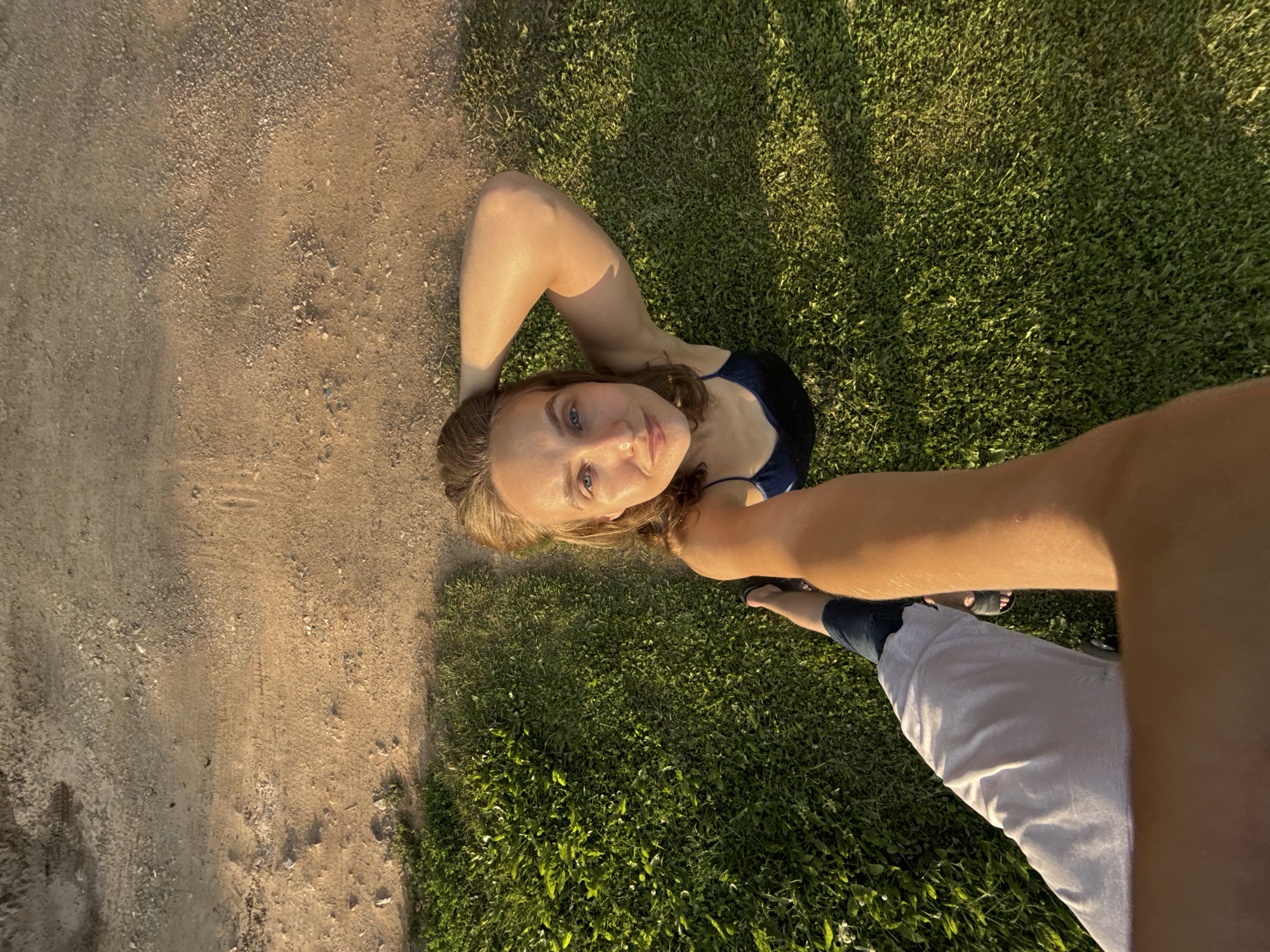 A high-angle, first-person perspective selfie is taken outdoors, with the camera held by a person whose arm extends into the lower left of the frame. The arm is covered by a light-colored sleeve and the person's legs are visible, wearing blue jeans and dark sandals. The foreground shows a patch of lush green grass, illuminated by warm, golden sunlight. Beyond the grass, a dry, dirt path with scattered gravel stretches across the upper portion of the image. The main subject, a young woman, is positioned in the upper right quadrant of the frame, looking directly at the camera. She has light brown hair pulled back and is wearing a dark blue tank top. Her left arm is raised, with her hand resting on her head. The scene appears to be in a park or a similar outdoor public space. The lighting suggests it is late afternoon or early evening.