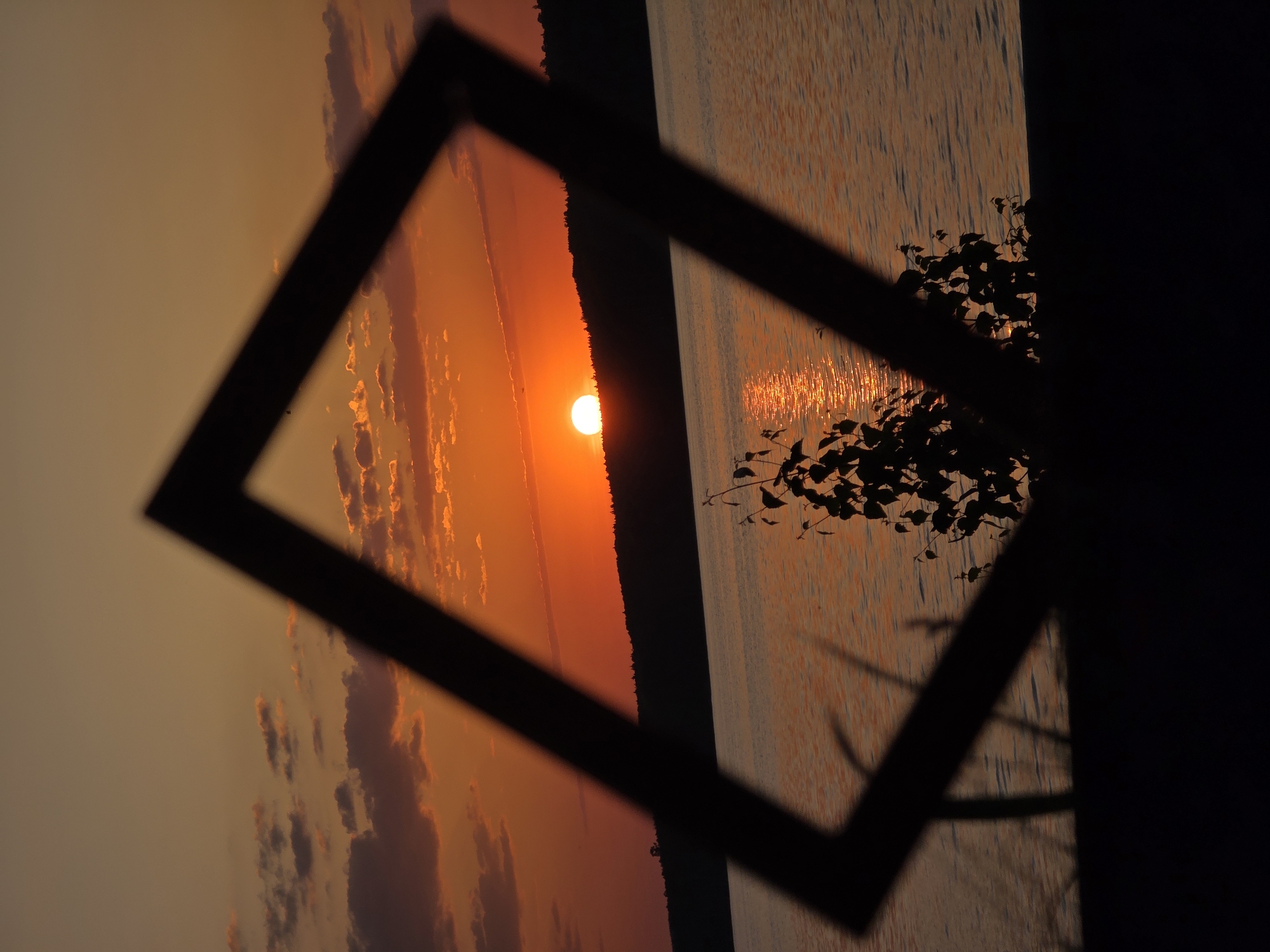 The image depicts a sunset, likely over a body of water, framed by a black square object. The lower right area is heavily obscured by shadow, but appears to have some foliage present. On the left is the orange and red sunset, with visible clouds and sun. The water surface is reflecting the light, seen within the frame. The general location is outdoors. A specific location within the city of Chaikovsky, Russia, cannot be identified without additional details.