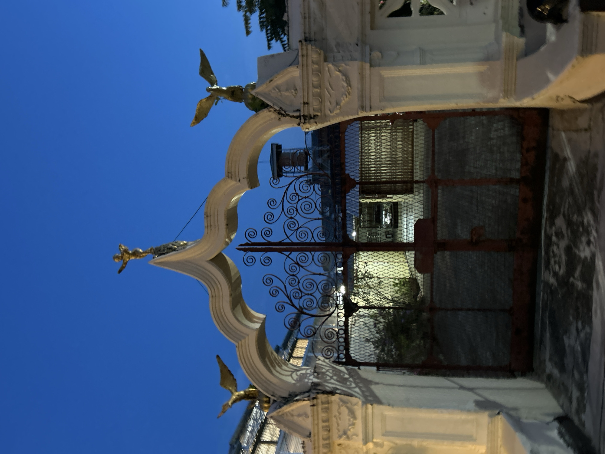 The image shows a wrought-iron gate topped by a decorative white archway, at dusk in Phuket City Municipality, Thailand. The archway is adorned with three golden statues: one of a winged figure on the apex and two winged creatures, possibly eagles, on either side. The gate is made of rusty red metal with intricate scrolling patterns. Beyond the gate, a dimly lit courtyard is visible, with some vegetation and structures in the background. The sky is a deep blue, indicating the twilight hour. The overall scene suggests an entrance to a property with some historical or artistic significance.