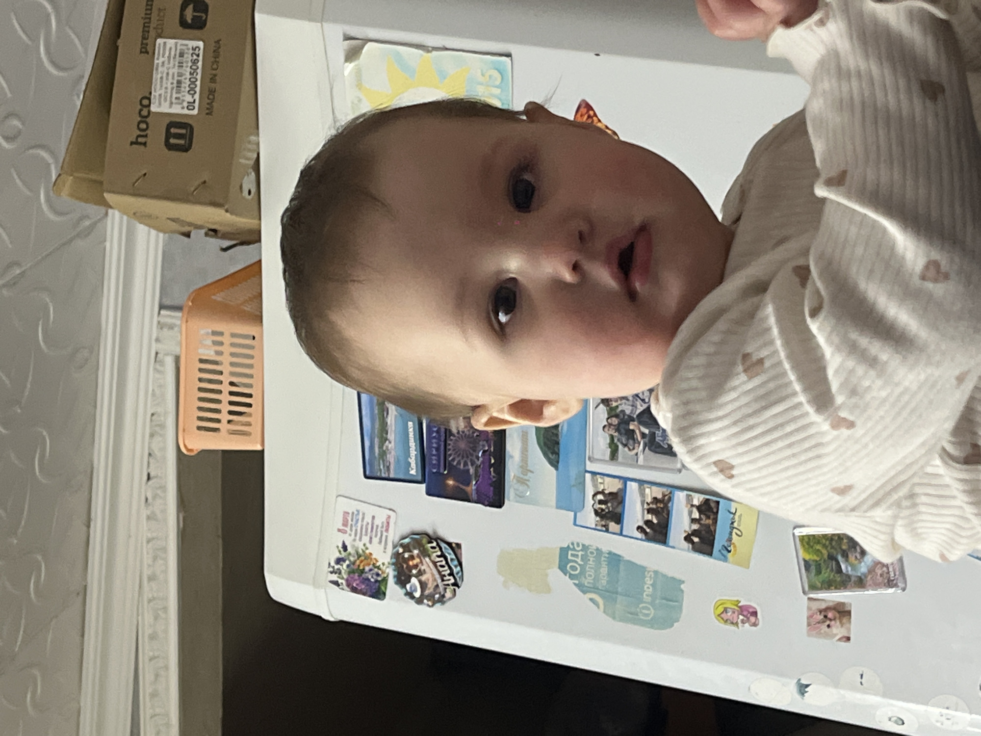 A baby in a white sweater is standing near a white refrigerator covered in magnets. The refrigerator is located in Novopokrovskaya, Russia. There is a box labeled "hoco" on top of the refrigerator. An orange basket hangs near the top of the refrigerator.