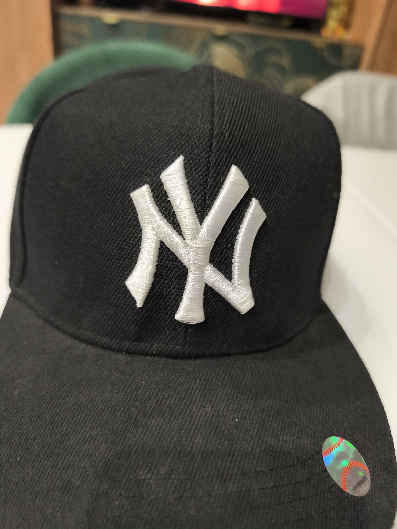 A close-up shot features a black baseball cap adorned with a white "NY" logo on the front. A holographic sticker is affixed to the right side of the cap's brim. The background is blurry and indistinct, comprising shades of green, white, and brown. The city of Moscow, Russia is not discernible from the image.