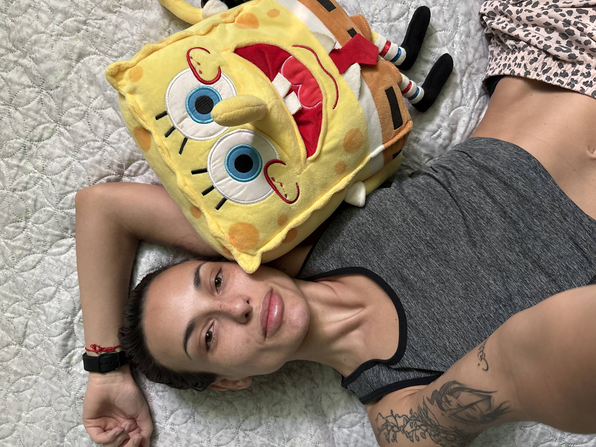 A woman lies on a white quilted bed. She wears a gray tank top, and part of her patterned pajama pants are visible in the upper right corner. A SpongeBob SquarePants plush toy rests on her head. Her left arm is extended outward, with a black smartwatch and red string bracelet visible on her wrist. A tattoo is seen on her right forearm. The woman's face is toward the camera, and she is looking into the lens. The background is a simple white quilted bedspread. The location is not discernible as Pleven, Bulgaria.