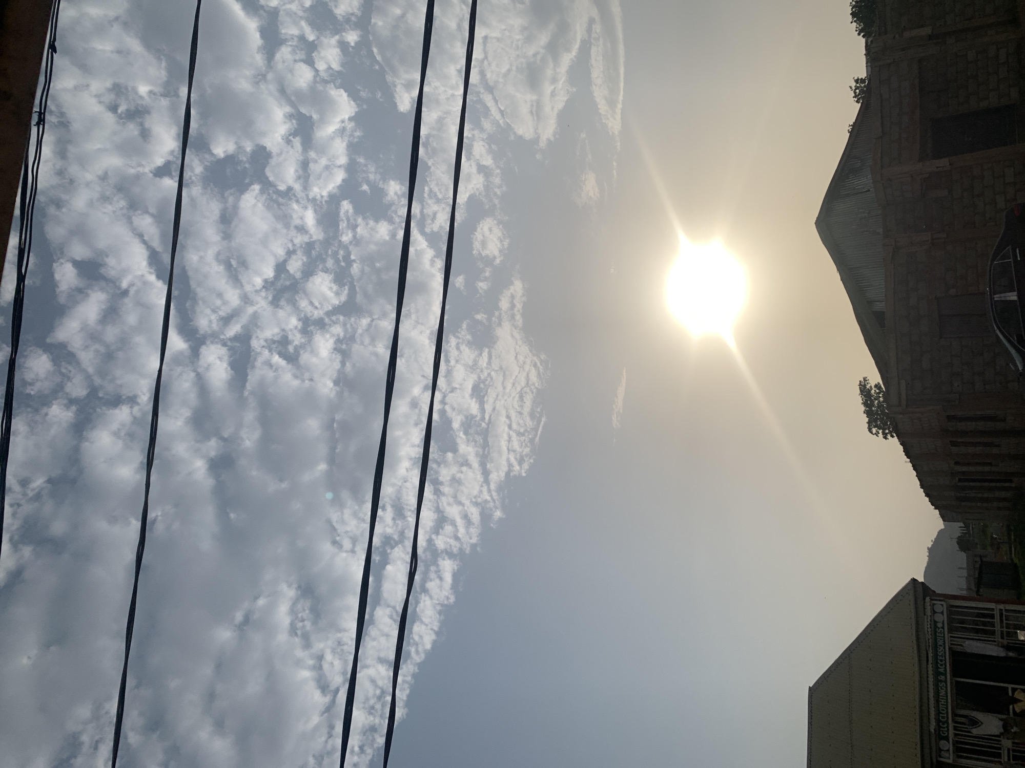 The image depicts a daytime scene in Sabojibi, Nigeria. The sky is predominantly filled with altocumulus clouds, transitioning from dense formations to a clearer, hazy expanse obscuring the sun. Several power lines stretch across the left side of the frame. On the right, parts of two buildings are visible: one under construction with exposed brickwork and a corrugated metal roof, and another with a sign reading "GLC CLOTHINGS & ACCESSORIES." A vehicle is partially visible next to the building under construction. The environment appears dusty or hazy.