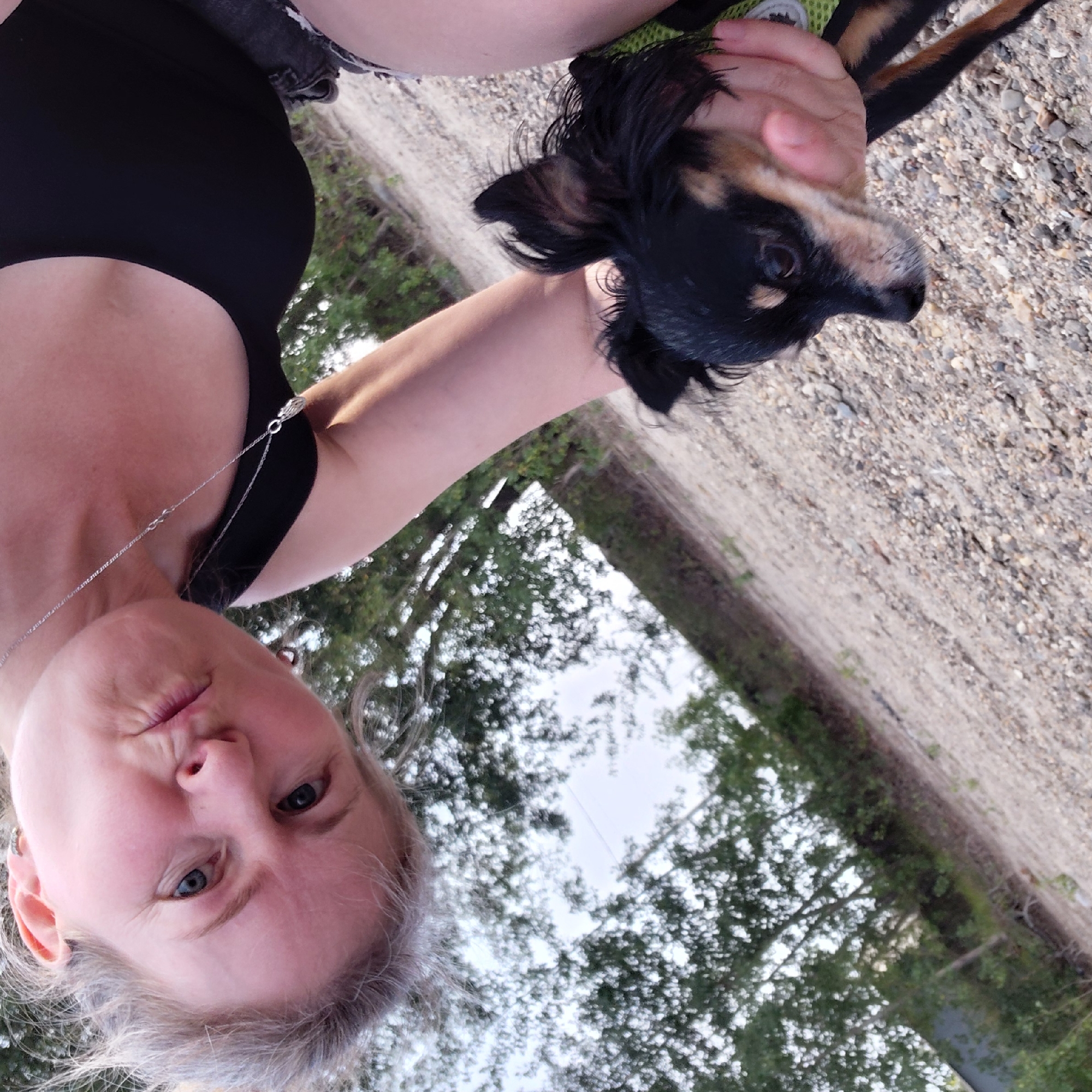 A woman with blonde hair is taking a selfie outdoors, with a small black and tan dog. The woman is looking at the camera with her lips pursed. The dog is being held by the woman, with its head and upper body visible. The background is a gravelly surface with trees and foliage. The image appears to be taken during daylight. The location is not specified as Novosibirsk, Russia.
