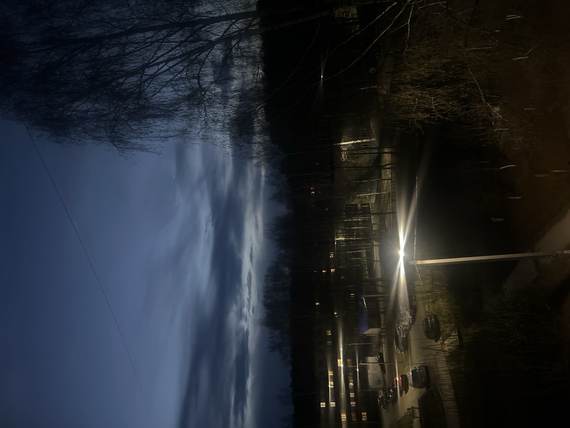 The image depicts an urban scene in Chaikovsky, Russia, at dusk or night. On the right of the image is a paved area and a street with parked cars. A street lamp illuminates the scene. Behind the street, several buildings with illuminated windows can be seen. A dark line of trees separates the buildings from the sky, which is filled with dark clouds. Bare tree branches are visible in the upper part of the image. A power line stretches across the left side of the sky.