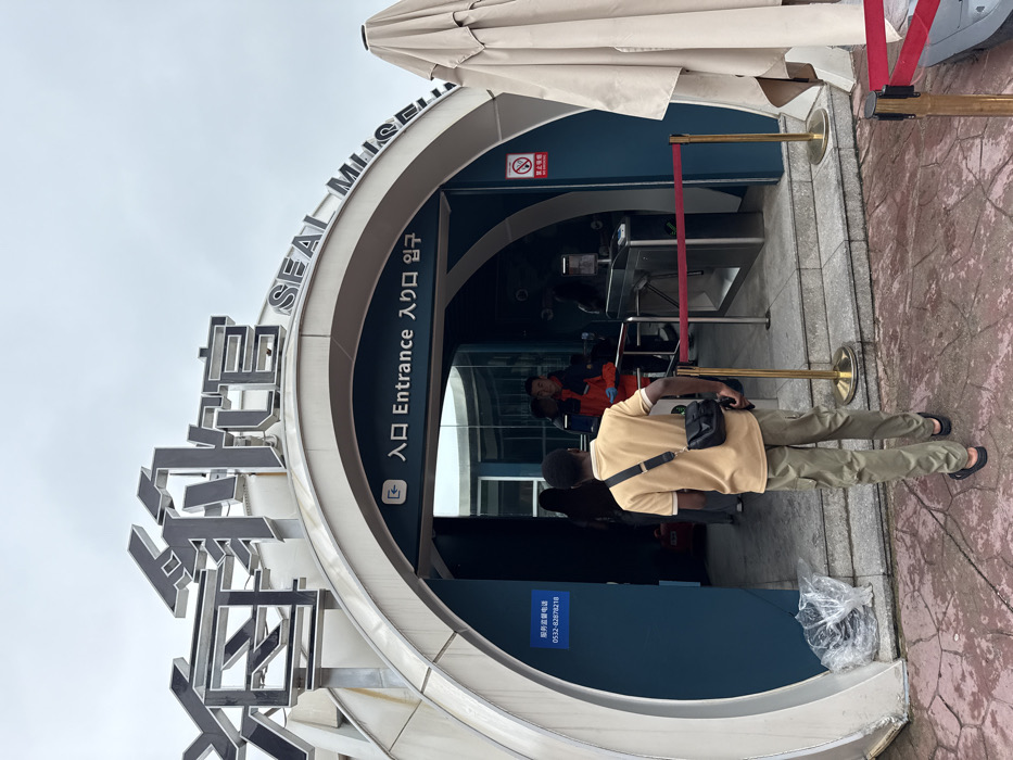 The image depicts the entrance to the Seal Museum (海豹馆 SEAL MUSEUM) located in Shinan District, China. The building features a modern, arched facade with metallic grey exterior panels and a deep teal interior section at the entrance. Above the entryway, large, dark grey Chinese characters for "海豹馆" are prominently displayed, followed by "SEAL MUSEUM" in English. Directly above the inner glass doors, a smaller sign in multiple languages reads "入口 Entrance 入口 입구".
In the foreground, a male individual with dark skin, wearing a light tan polo shirt, olive green cargo pants, and black sandals, walks away from the viewer towards the museum entrance. He carries a black shoulder bag. Inside the entrance, an automated stainless steel turnstile system is visible, preceded by red velvet ropes on brass stanchions, forming a queue line. Other individuals are visible beyond the turnstiles; one person wears a red and orange jacket. A blue sign on the left interior wall displays a Chinese service supervision phone number: "0532-82878218". On the right interior wall, a red and white "no smoking" sign is affixed. To the far right of the entrance, a large, beige, folded outdoor umbrella is partially visible. The ground is paved with reddish-brown, irregularly shaped flagstones under an overcast sky. A clear plastic bag containing trash is positioned near the base of the left arch.