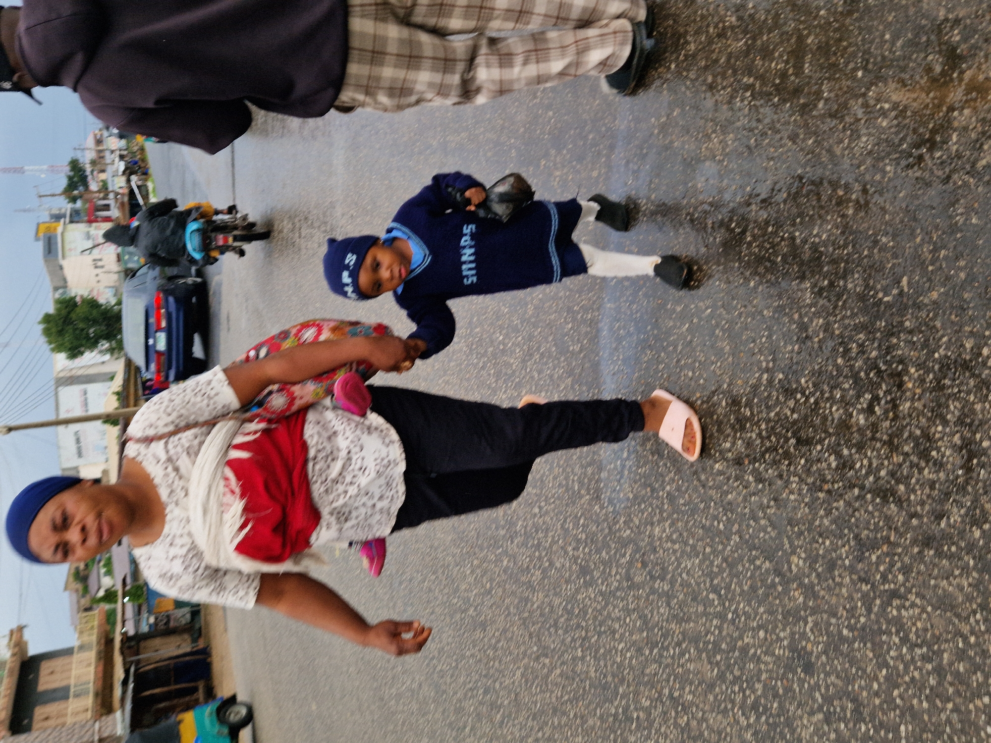 The image depicts an overcast, wet street scene in Kabong, Nigeria. A woman wearing a blue headscarf and a white patterned shirt is walking, carrying a child on her back wrapped in a red cloth and a white fabric. She is holding the hand of a small child walking beside her, who is dressed in a blue sweater, blue hat, white leggings, and black shoes. The child is also carrying a dark object in their hand. Another person, a male wearing a dark hoodie and plaid pants, is visible in the foreground to the right. In the background, a blue car is driving on the road, and a motorcycle with a rider wearing a dark jacket is also present. Various buildings and structures line the street, some appearing to be shops. The road surface is wet and reflects the ambient light, indicating recent or ongoing rain. A portion of a yellow and turquoise three-wheeled vehicle is visible on the left side of the image.