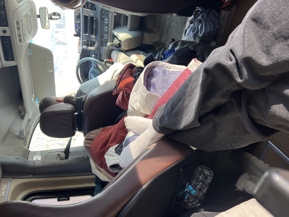 The image depicts the interior of a Volkswagen multi-passenger vehicle, likely a van, in Samolet, Russia, during daylight hours. The perspective is from a rear passenger seat, looking forward. In the immediate foreground, the legs of a person are visible, clad in dark grey pants and white crew socks, propped up over a pile of garments. This pile, consisting of various jackets in colors such as red, white, purple, and blue, occupies the space between the front seats. The driver, a male wearing a light-colored long-sleeved shirt, is visible from the rear, hands on the steering wheel bearing the Volkswagen emblem. A second passenger's head is partially visible on the far right. The vehicle's dashboard features a manual gear shifter and an infotainment unit. An air freshener hangs from the rearview mirror. On the left, a brown leather-like seat has a mesh pocket containing a blue object. A white furry slipper or boot is on the floor near the foreground passenger's feet. Through the front windshield, a brightly illuminated outdoor environment with sparse trees and a light-colored ground, possibly snow, is discernible.