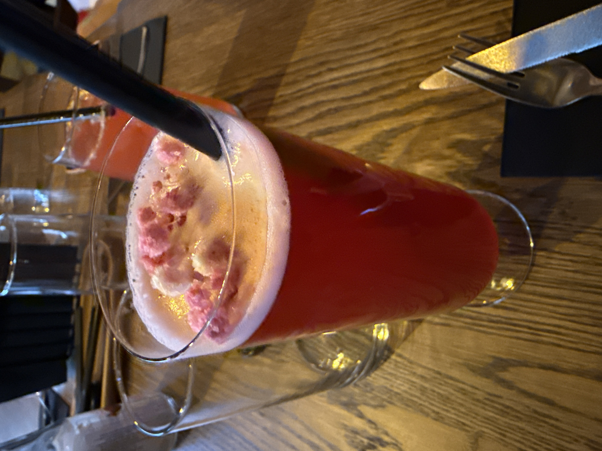 The image shows a close-up of a red beverage in a tall glass, topped with foam and small pink pieces, likely a garnish. A black straw is visible in the drink. To the left of the main drink, another glass is partially visible. In the background, a second drink with a similar red color is also visible, with a straw. The drinks are placed on a wooden table. To the right of the main drink, a fork and knife are laid out on a dark surface, suggesting a dining setting. The lighting is warm and casts shadows on the table. The scene appears to be indoors, possibly in a restaurant or bar. No specific location in Sofia, Bulgaria can be identified from the image alone.