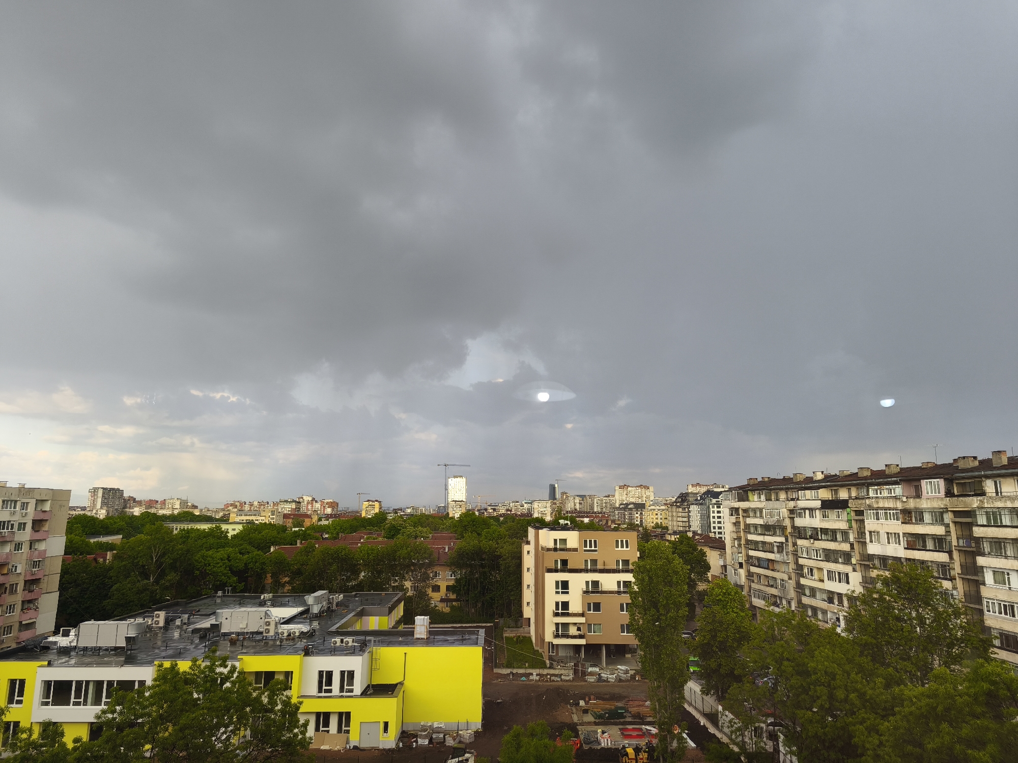 The image shows a view over a residential area in Sofia, Bulgaria, under a cloudy, overcast sky. In the foreground, there are several buildings, including a yellow and white low-rise structure, likely a school or kindergarten. Adjacent to it is a newly constructed apartment building in earth tones with exposed construction materials and equipment around it. To the right stands a multi-story apartment building, typical of post-war socialist architecture, with trees partially obscuring its façade. In the distance, the skyline of Sofia is visible with several modern high-rise buildings and cranes. The lighting suggests a dull day with the overcast sky casting a flat light over the scene. There is no evidence of any people visible in this scene.