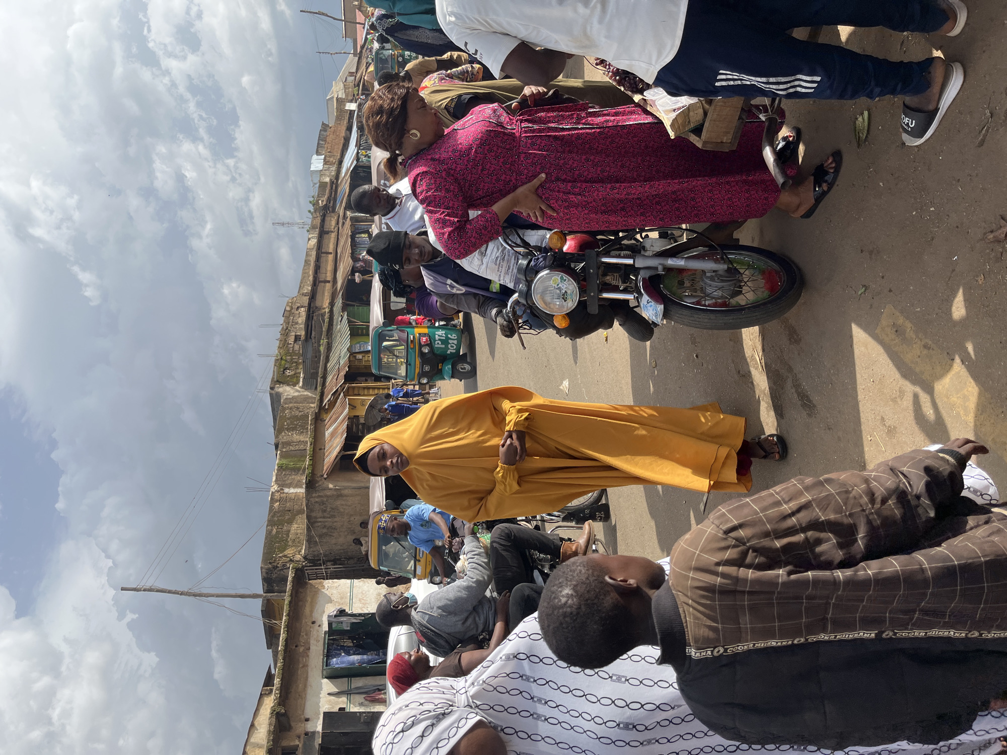 The image depicts a bustling street scene in Rafin Par, Nigeria, likely a marketplace given the presence of various stalls and vehicles. In the foreground, a young boy wearing a brown and black checkered jacket faces away from the camera. To his right, a woman in a bright yellow hijab and long dress stands looking towards the viewer, her hands clasped in front of her.
Further into the scene, a woman in a vibrant pink dress is leaning forward, her right hand on her hip, looking down. She is standing next to a motorcycle. A man in a black beanie is seated on the motorcycle, appearing to be the driver. Behind them, a man in a white shirt is looking towards the motorcycle. Several other individuals are visible in the background, along with three auto-rickshaws, suggesting active commerce and transportation. The buildings lining the street appear to be modest, with some showing signs of wear. The sky is cloudy, with patches of sunlight breaking through. The overall impression is one of a lively and active urban environment.