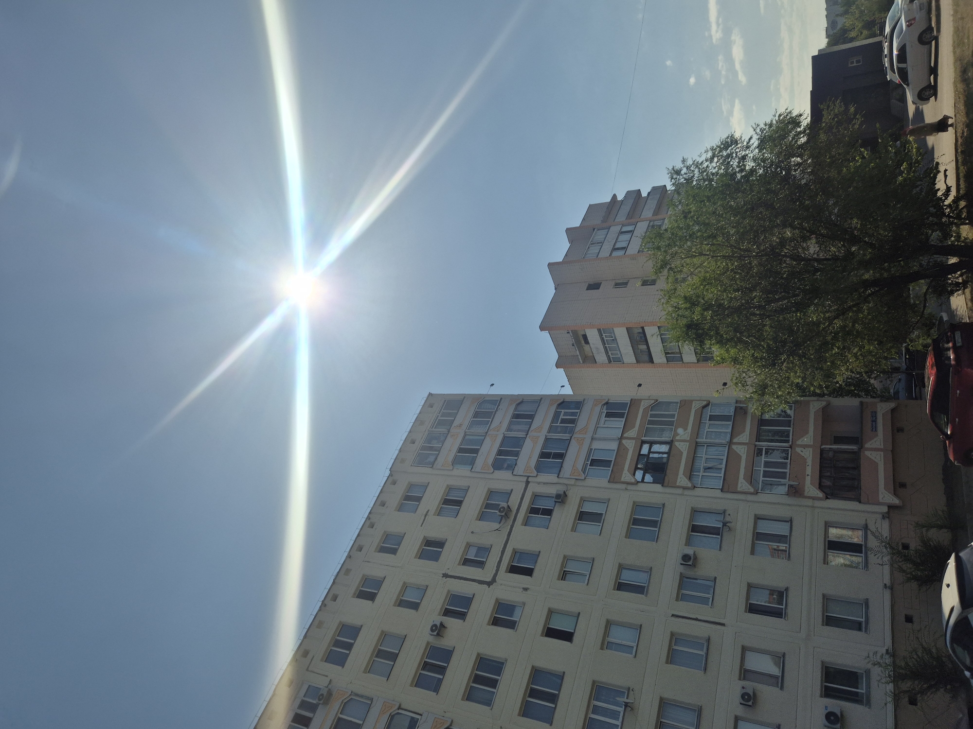The image shows a bright, sunny day with a clear blue sky. The sun is high in the sky and is creating a strong lens flare effect. In the foreground, there are two apartment buildings. The building on the left is taller and wider, with a light-colored facade and numerous windows. The building on the right is shorter and narrower, with a light beige facade and balconies. A large, green tree is positioned between the two buildings, partially obscuring the right building. In the lower right corner, there is a sidewalk with a few parked cars and a person walking. The scene appears to be in a residential area of Kokshetau, Kazakhstan, given the architectural style of the apartment buildings and the presence of parked vehicles typical of urban environments.