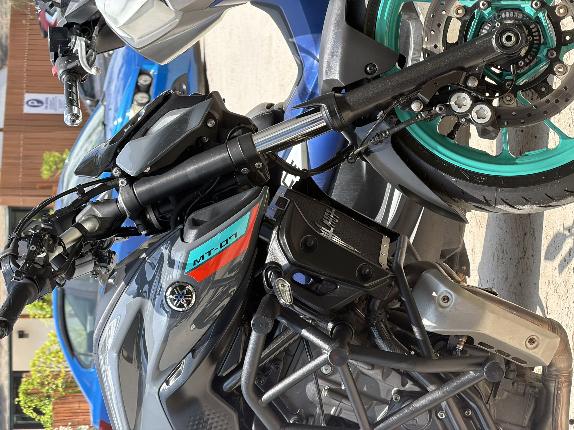 A grey Yamaha MT-07 motorcycle is the primary subject, viewed from a high angle, capturing its front and middle sections. The motorcycle features bright teal/green wheels, notably visible on the front wheel's brake disc and caliper. It has aftermarket black crash bars protecting the engine area, and a grey heat shield over part of the exhaust system. The handlebars, front forks, and various black components are clearly visible.
In the background, a blue vehicle, likely a car, is partially visible, parked behind the motorcycle. Further back, parts of buildings or walls with a textured surface, and some green foliage from potted plants or small trees, suggest an outdoor urban or suburban setting. The bright light and distinct shadows indicate it is daytime, likely midday or early afternoon, with sunny weather.
Visible text includes "MT-07" in teal and red on the side panel of the motorcycle, and the Yamaha logo on the fuel tank. A partial "P" symbol is seen on a white sign in the background, suggesting a parking area. Faint, unreadable text is present on the tire sidewall and brake caliper. The word "ALPHA" is partially visible on a black panel above the crash bars near the engine. No specific activity is taking place; the motorcycle appears to be parked. The overall scene depicts a well-maintained motorcycle in an outdoor setting in Choeng Thale, Thailand.