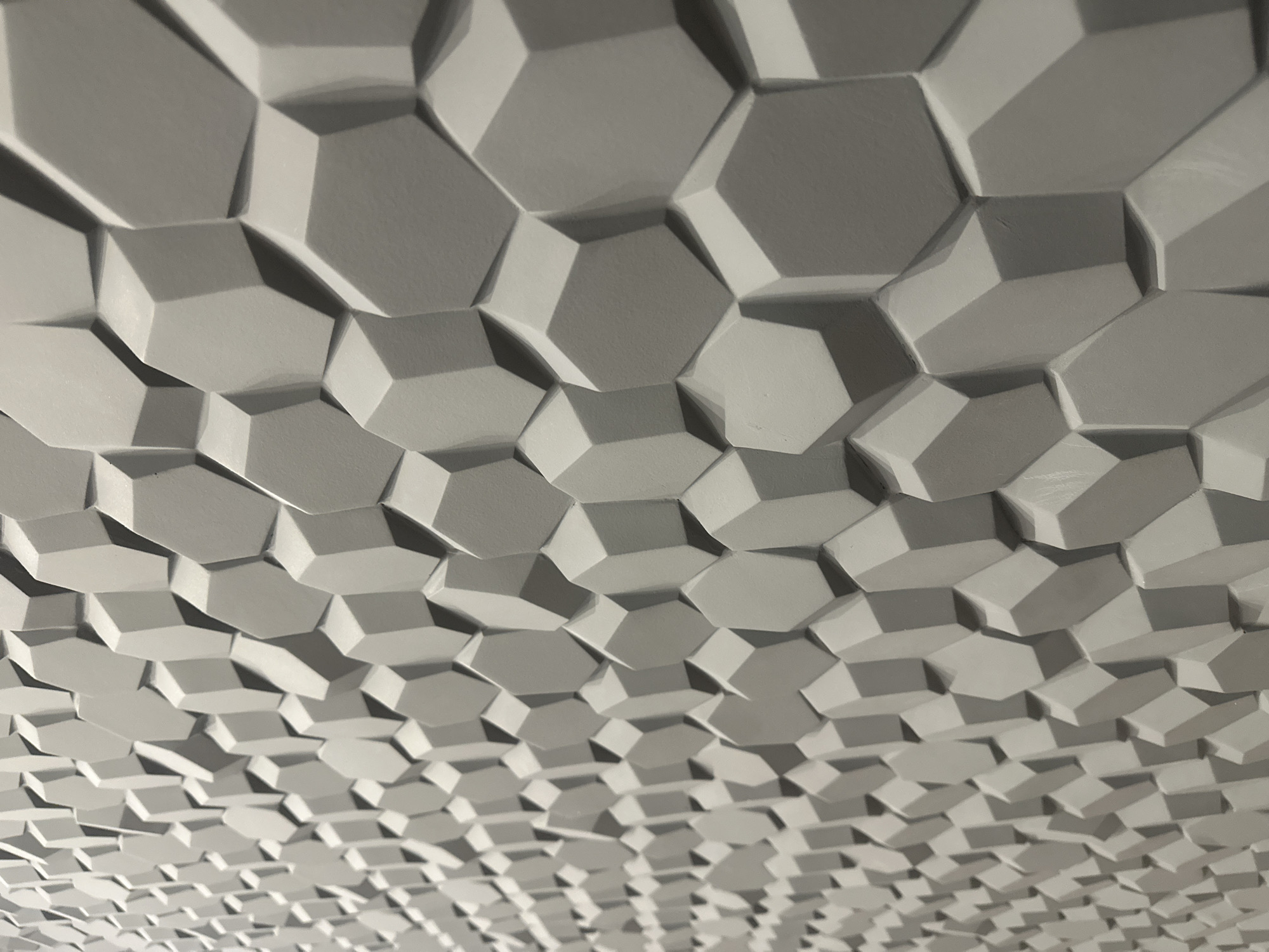 The image presents a detailed, oblique view of a three-dimensional textured wall surface. The surface is uniformly composed of numerous interlocking, raised polygonal shapes, predominantly irregular hexagons or pentagons, finished in a consistent matte light grey or off-white color. These geometric modules vary slightly in their projection from the wall, creating a dynamic, faceted relief. Illumination from an unidentifiable source, likely artificial, casts distinct shadows on the left and lower edges of the raised elements, enhancing their depth and three-dimensionality. The perspective provides sharp focus on the larger, more prominent polygons in the mid-to-right foreground, while the pattern recedes towards the upper left, appearing smaller and slightly out of focus. No people, objects, or environmental details beyond the wall surface itself are discernible. The specific location in the city of Izhevsk, Russia, cannot be determined from the provided image.