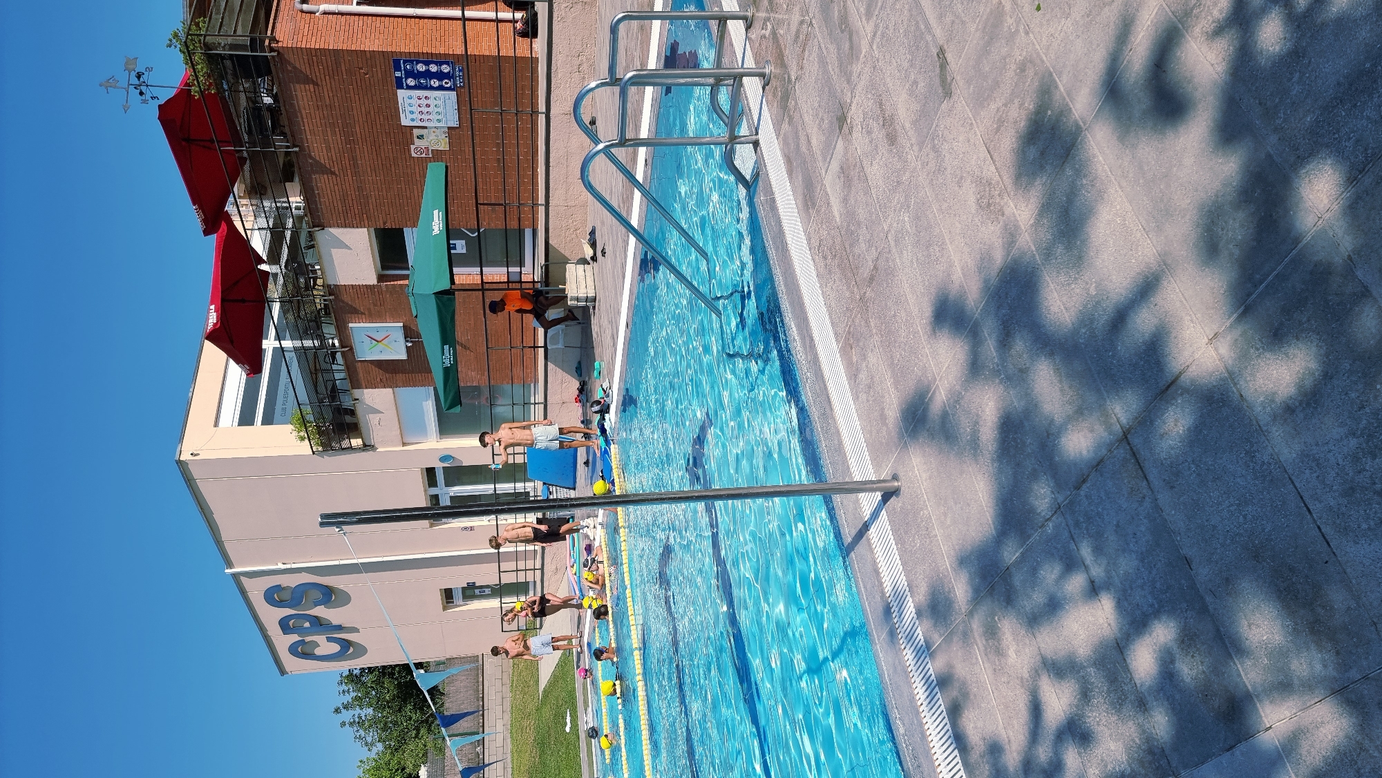 The image shows a swimming pool area at Club Poliesportiu Sant Joan Les Fonts, located in Sant Joan Les Fonts, Spain. The facility has a light beige building with the blue letters "CPS" prominently displayed, and a red-tiled section. Red and green umbrellas are set up on a patio area. Several lanes are marked in the blue swimming pool with yellow lane markers. In and around the pool, there are several individuals. In the pool, some are swimming or training, while others are in shallow water. On the poolside, a person in an orange shirt is seated on concrete blocks. Another person in light-colored shorts is standing near the edge of the pool, holding a clear bottle. The ground around the pool is paved with gray tiles, and tree shadows are cast across the surface. The sky is clear and blue, indicating a sunny day.