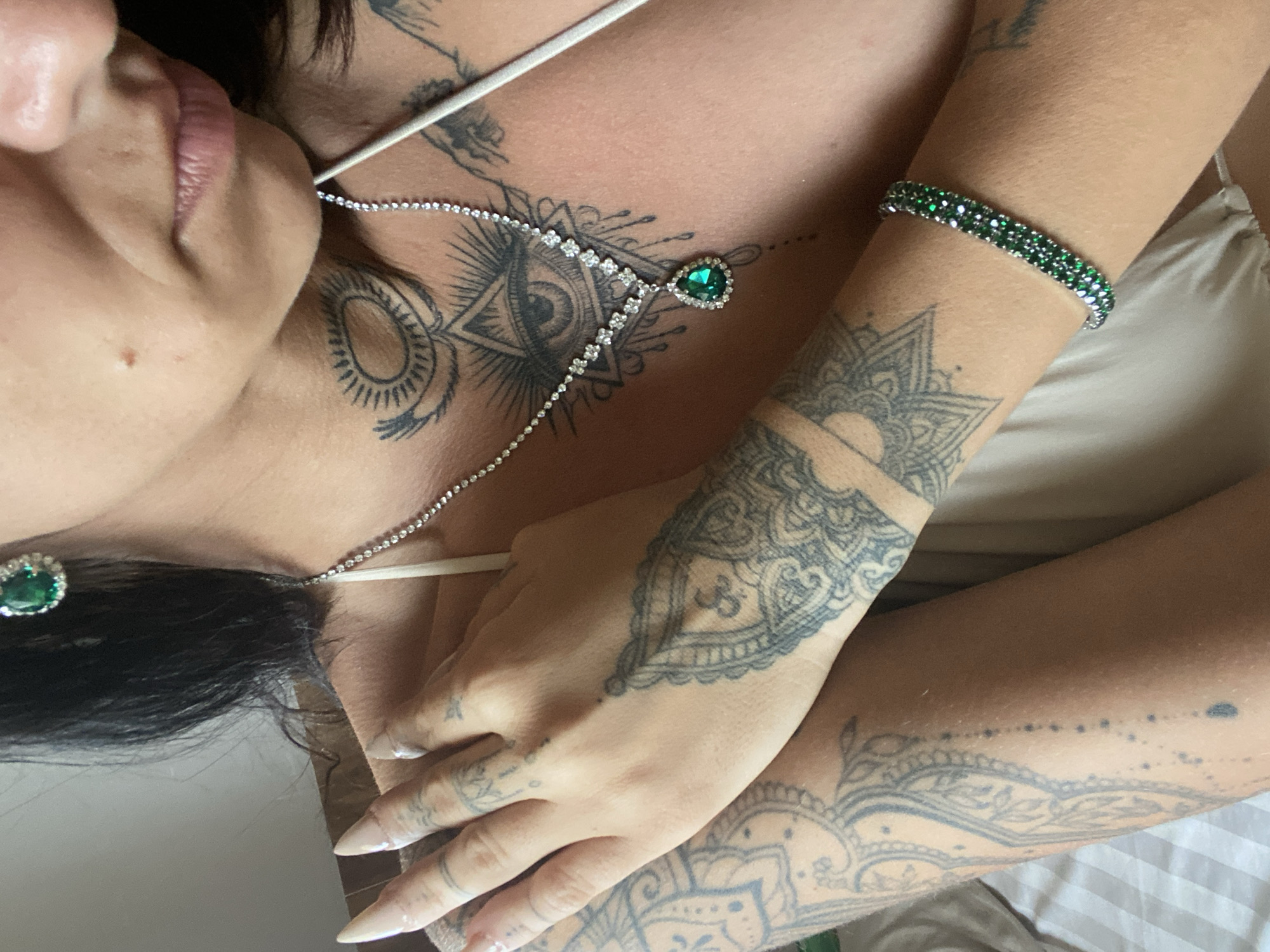 The image shows a close-up of a person's upper chest, neck, and arm, adorned with jewelry and tattoos. The person is wearing a delicate silver necklace with a pear-shaped green gemstone pendant, surrounded by small clear stones. They also have matching green gemstone earrings and a green gemstone bracelet. The tattoos are intricate, featuring a large eye with rays in the center of the chest, a sun-like design on the neck, and various patterns on the arm. The skin tone suggests an outdoor setting, possibly a beach or resort. The background is a soft focus of light and shadow, hinting at a bright environment. No specific location in Mũi Né, Vietnam can be determined from the visual information provided.
