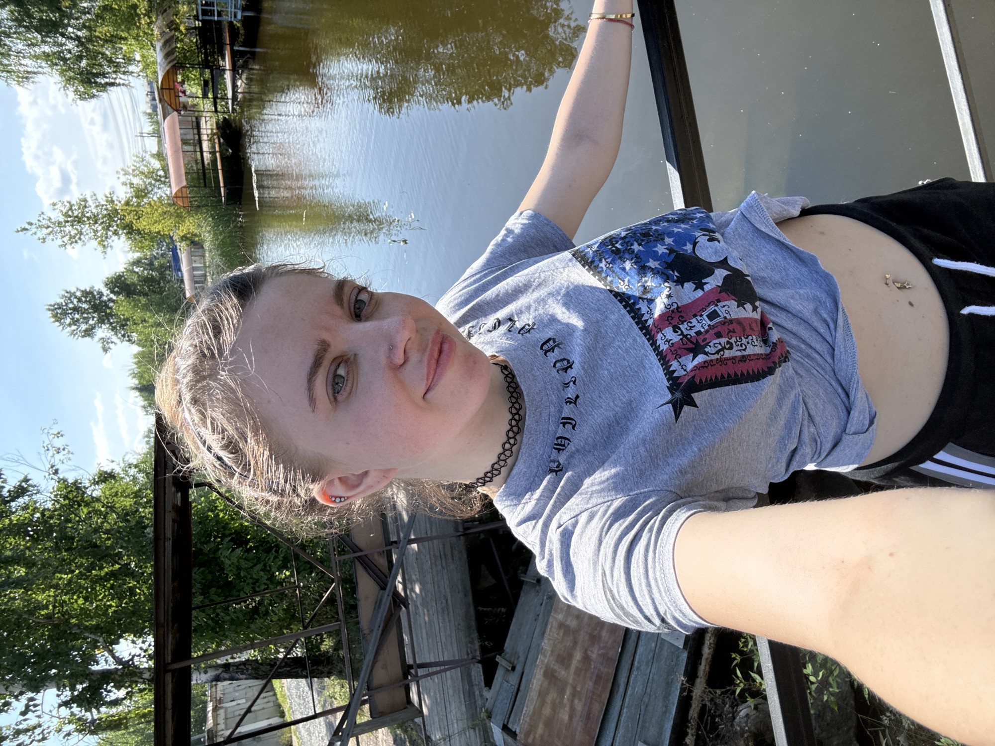 A young woman with blonde hair pulled back and tied into a ponytail takes a selfie outdoors on a sunny day. She is wearing a grey t-shirt with an American flag design on the front and black shorts. She has a black choker necklace and a belly button piercing. She is standing on a bridge or boardwalk overlooking a body of water. In the background, there are trees, buildings, and a pier with covered seating areas. The overall scene is casual and appears to be in a recreational area.