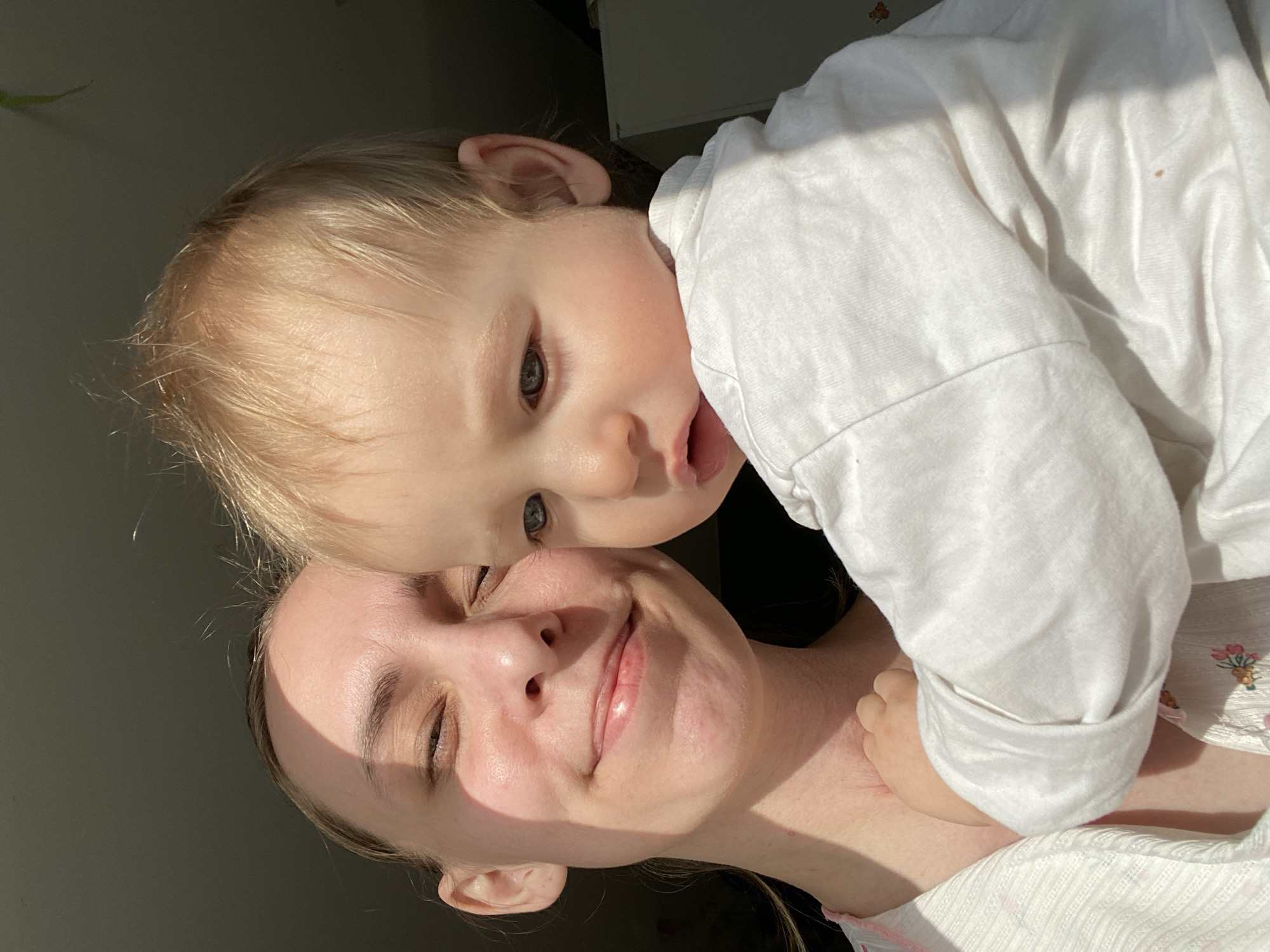 A woman is holding a toddler. The woman has fair skin, and is fair-haired. The toddler is wearing a white shirt. The image location appears to be indoors, lit by sunlight. The location cannot be confirmed to be Chaikovsky, Russia.