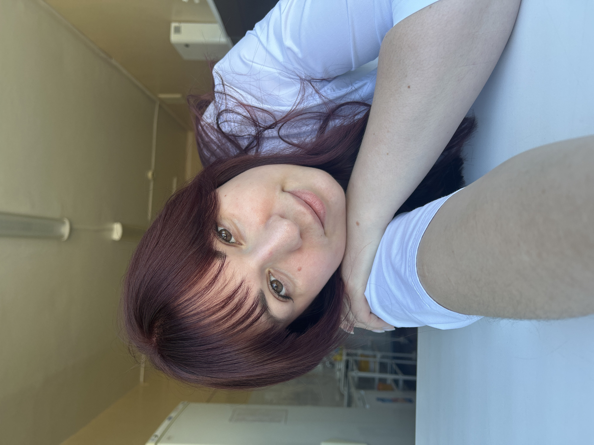 A woman with auburn hair is lying on a white surface. She is wearing a white t-shirt. The location appears to be indoors, with a cream-colored wall visible in the background. The scene is cropped tightly around her face and upper body, with a blurred view of a structure with some visible elements resembling scaffolding or shelving through a window. No definitive elements link the location to Фоки, Russia.