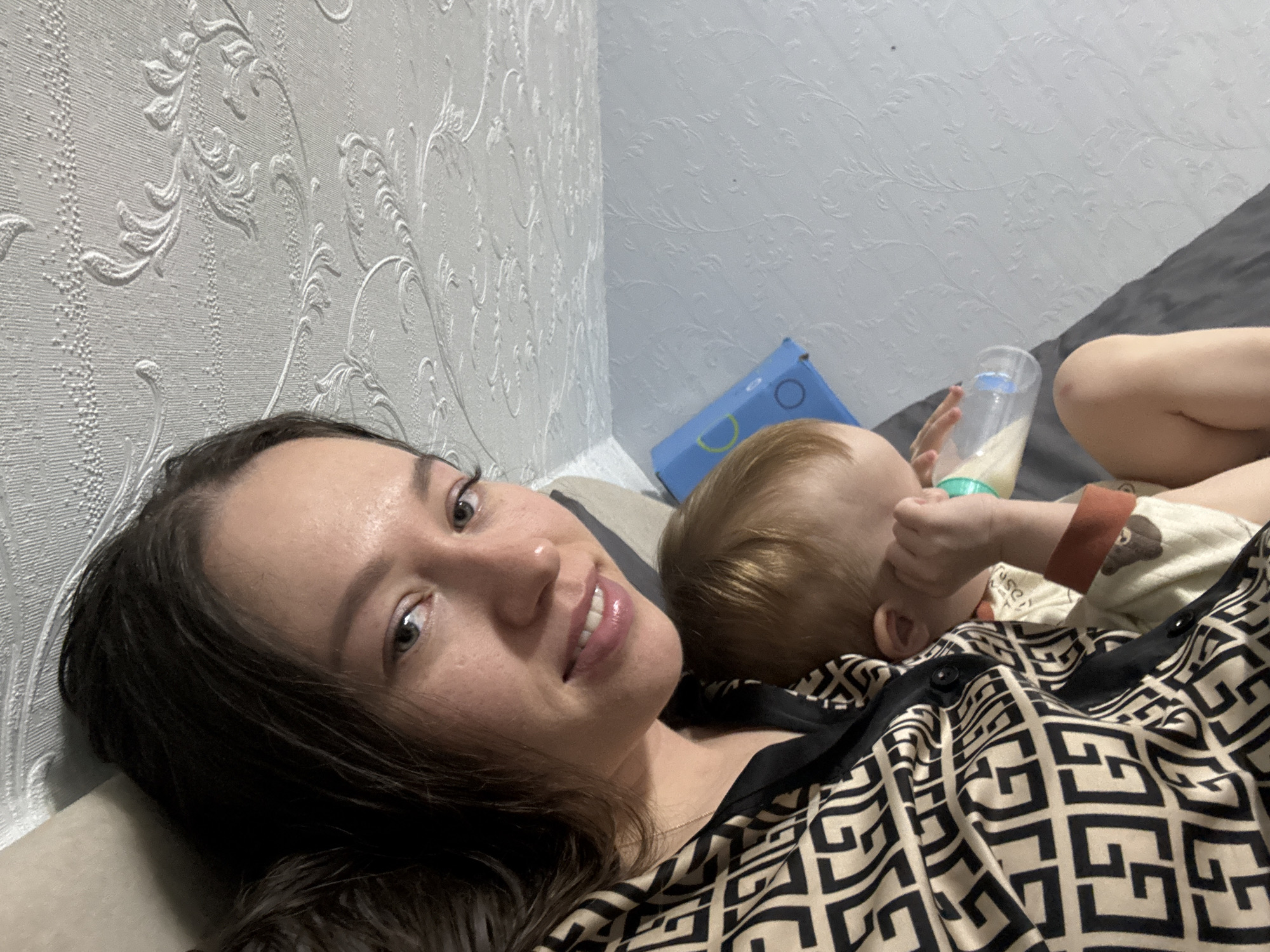 A fair-skinned woman with brown hair lies on a bed, angled towards the camera, in a room with light gray patterned wallpaper and gray bedding. She is wearing a black and tan patterned top. A baby with light blonde hair is also on the bed, drinking from a bottle. A blue box is visible in the background. The location is Ферма, Russia.