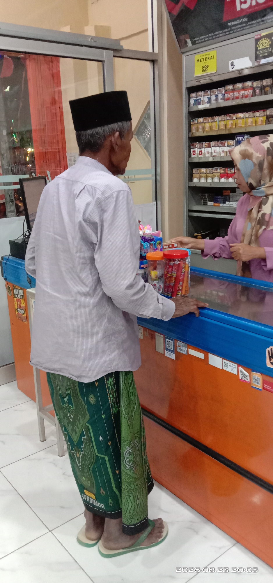 A man wearing a black peci hat and a light gray pinstripe long-sleeved shirt is standing at a counter, facing away from the camera. He is also wearing a green patterned sarong. He is interacting with a woman wearing a pink long-sleeved blouse and a patterned hijab. The woman is reaching towards the counter, which is filled with various items, including cigarettes displayed on shelves behind her. The counter itself has a blue edge and an orange base, with several stickers and QR codes on its side. In the foreground, there are clear plastic containers filled with what appear to be candies or snacks. The man is wearing flip-flops. The environment appears to be an indoor store, possibly a convenience store or minimarket, with tiled flooring. A date stamp at the bottom right indicates "2026.08.23 20:05". The location is identified as Pakong, Indonesia.