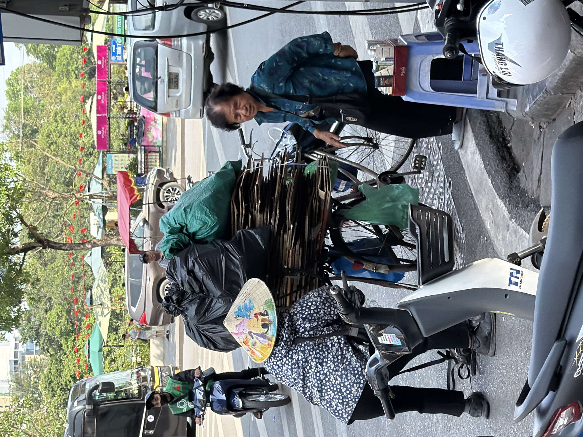 This image captures a vibrant street scene in Hà Nội, Vietnam, during daytime under clear, sunny weather. The setting appears to be a busy urban street or sidewalk, indicative of daily life and commerce.
In the foreground, two individuals are prominent. To the right, an elderly woman in a patterned blue-green long-sleeved shirt and dark trousers stands next to a heavily laden bicycle. She is looking towards the viewer. Her bicycle is piled high with flattened cardboard or similar brown items, secured with ropes, and includes green fabric bags. Next to her, a small light blue plastic stool holds several cigarette packs, with "Ruby" and "Zippo" visible, and other small items.
To the left, a person is seated on a scooter, wearing a traditional Vietnamese conical hat (nón lá) adorned with colorful painted scenes. This scooter is also burdened with black trash bags and green plastic bags, suggesting they are collecting or selling goods. Another scooter, partly visible in the lower left, has a rider wearing a green uniform with the "Grab" logo, indicating a delivery or ride-hailing service. The scooter in front of the conical hat has "TRI HAM GROUP" printed on its side panel.
In the background, other vehicles including cars and more scooters are visible, confirming a bustling environment. A white car in the mid-ground has "329.94" on its license plate. Further back, street stalls with awnings and lush green trees line the street. A pink banner with "LỄ THẤT TRUYỀN" and a green sign with "TAXI" are visible above the cars. A white helmet with "THANG LO" is seen hanging on a scooter handle in the mid-right. The scene conveys a sense of industriousness and the typical street activities found in Vietnamese cities.