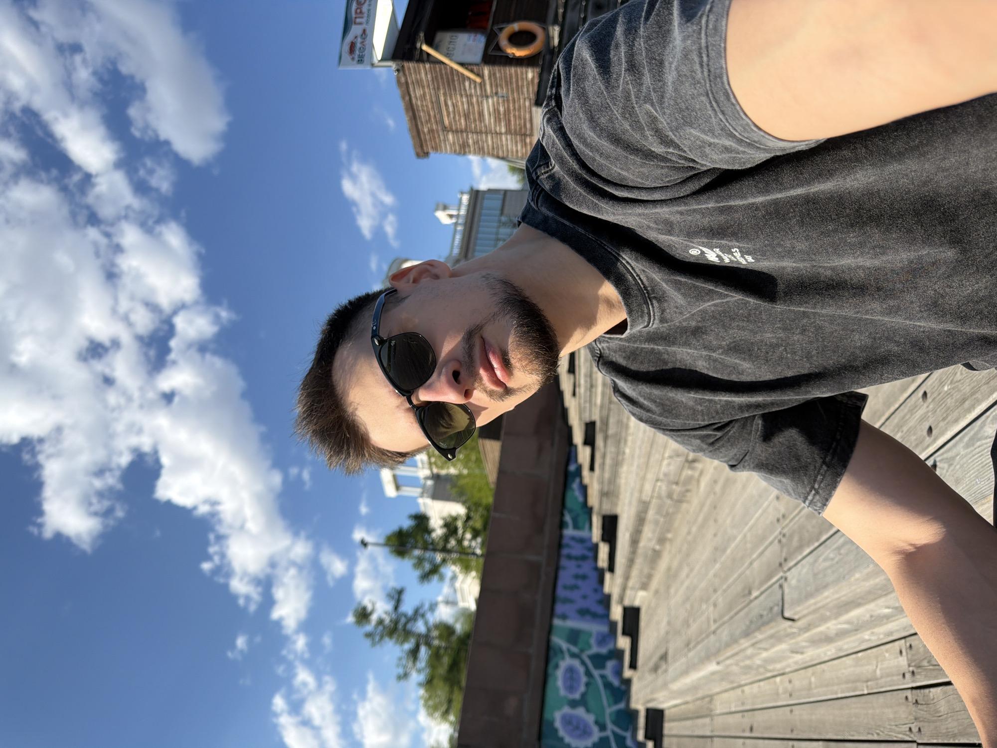 A man with a short beard and dark sunglasses is positioned in the foreground, taking a selfie. He wears a dark gray t-shirt. Behind him, to the left, is a blue sky with scattered white clouds. Below the sky, there are trees with green foliage. To the right of the man, there is a wooden boardwalk. Further in the background, there's a small building with a sign in Russian. It appears to be a summer kiosk named "Весла ПРО". The location seems to be in Krasnoyarsk, Russia.