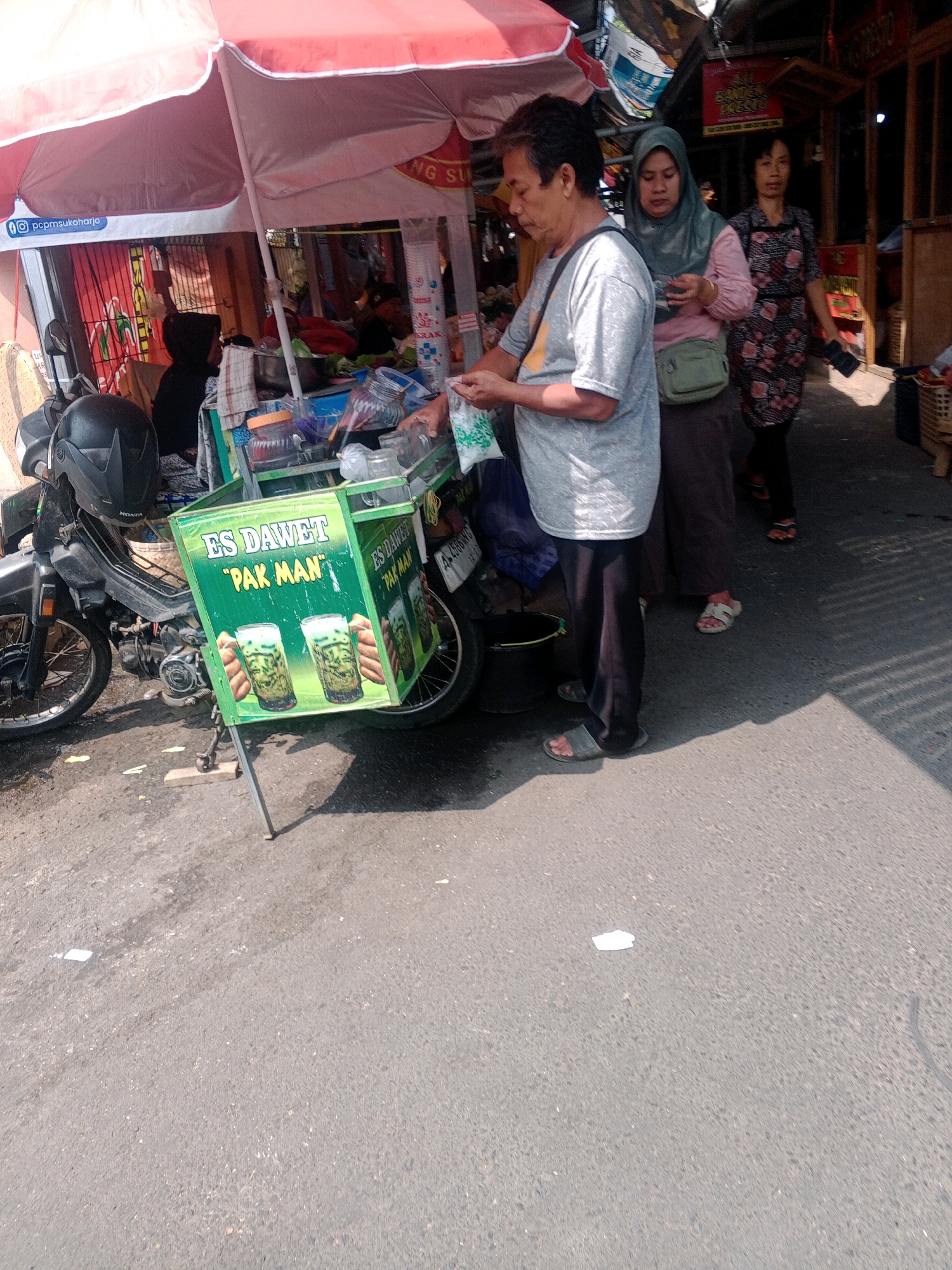 The image depicts a street scene in Wonogiri, Indonesia, likely a market area, with a focus on a beverage cart. A man in a grey t-shirt and dark pants is standing near the cart, holding a bag and interacting with the cart's contents. Behind him, two women are walking away. The woman in the foreground wears a green headscarf and a pink shirt, and the woman behind her is wearing a floral patterned dress. The cart itself is green with a sign that reads "ES DAWET PAK MAN" and features images of two drinks. A motorcycle is parked to the left of the cart, with a black helmet on it. The environment appears to be an outdoor market with stalls and awnings visible in the background. The ground is asphalt, and sunlight casts shadows.
