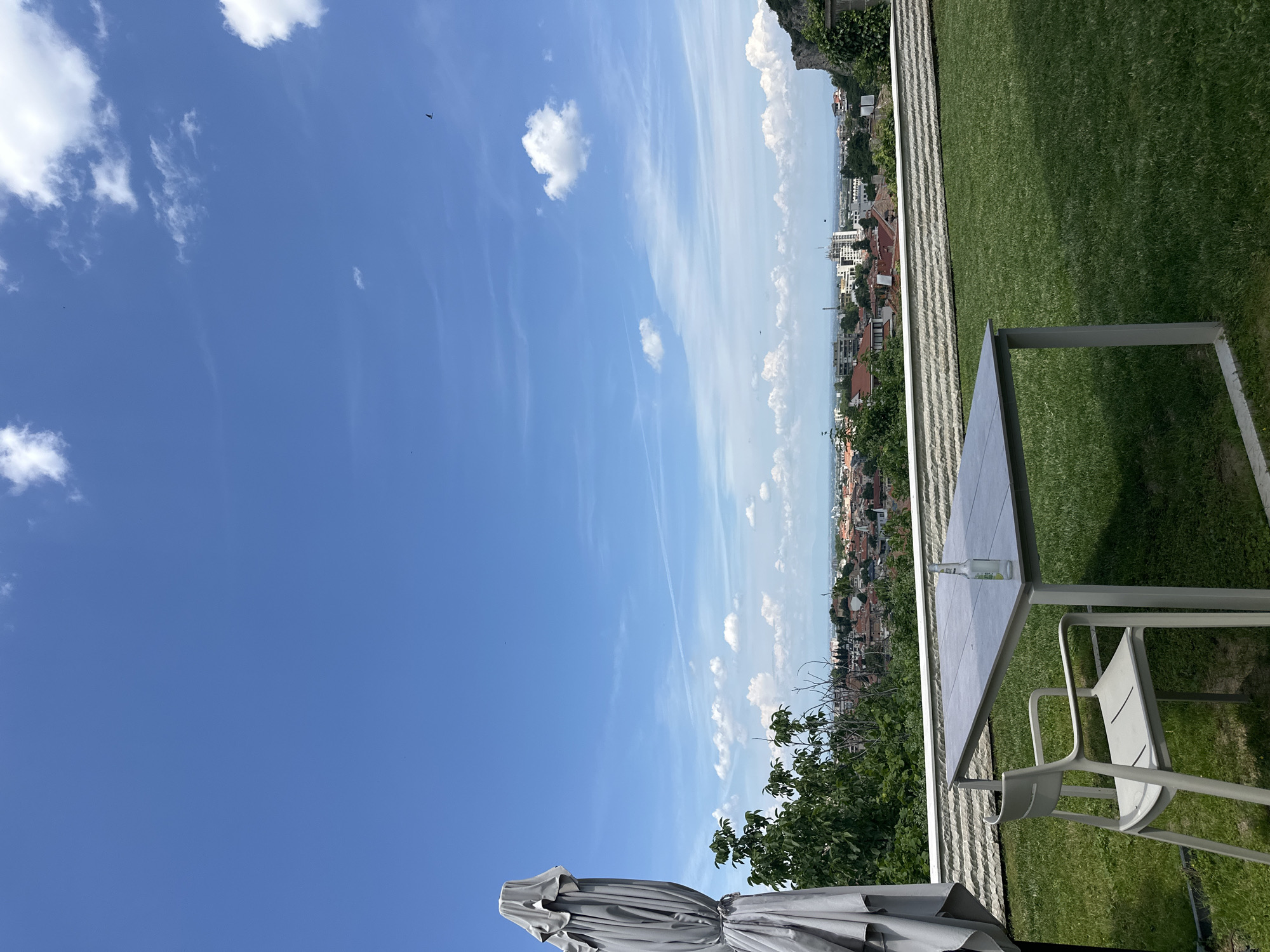 The image shows a high-angle view of an outdoor seating area with a panoramic view of Asenovgrad, Bulgaria. In the foreground, there is a grey outdoor table and a white chair situated on a well-maintained green lawn. A closed grey umbrella is visible on the left side of the frame. On the table, a single clear glass bottle with a light-colored liquid and a label is present.
The middle ground features a low, horizontal stone wall that separates the lawn from a densely populated urban area. The city of Asenovgrad unfolds beyond this wall, characterized by a collection of red-tiled roofs and various buildings, including some taller structures. Lush green trees are interspersed throughout the cityscape and on the slopes leading up to it.
The background is dominated by a vast, bright blue sky with scattered white clouds. The overall impression is one of a clear, sunny day, likely during the daytime, with a pleasant view from a high vantage point overlooking the city. There are no people visible in the image, and the scene appears to be static and undisturbed. The perspective suggests the photograph was taken from a balcony, terrace, or elevated garden area.
