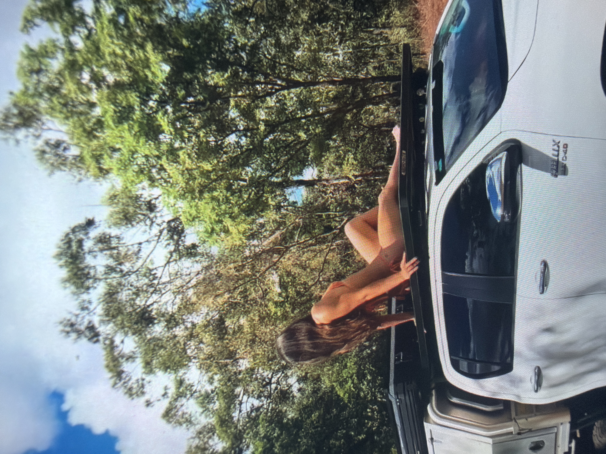 This image depicts an outdoor scene in Shumen, Bulgaria. A long-haired adult female, dressed in a light orange two-piece bikini, is positioned in a reclining pose on a black heavy-duty roof rack installed atop a white Toyota Hilux 4x4 D-4D pickup truck. Her left arm supports her upper body, and her right hand rests near her hip on the roof rack. The truck features dark-tinted windows, chrome door handles, a chrome side mirror cap, and a silver utility canopy at the rear. The background consists of dense green trees under a clear blue sky with white clouds visible in the upper left. The ground appears to be dry soil.