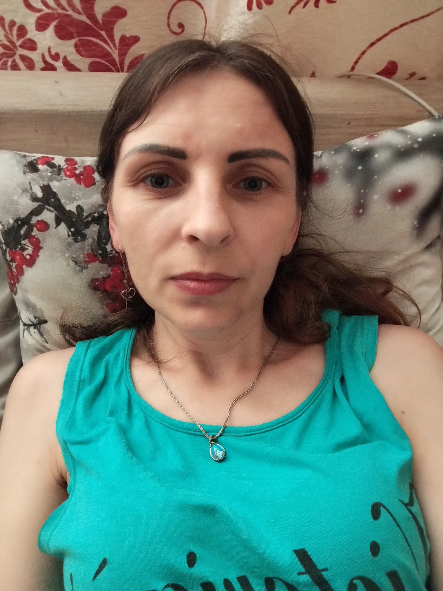 A selfie of a woman with brown hair and blue eyes is taken from a slightly high angle, with the woman lying down on a bed with patterned pillows. She is wearing a turquoise tank top and a silver necklace with a blue pendant. The background shows a headboard with a floral pattern. The woman's facial expression is neutral. The image does not contain any information that would allow for the determination of its location in Vinnytsia, Ukraine.