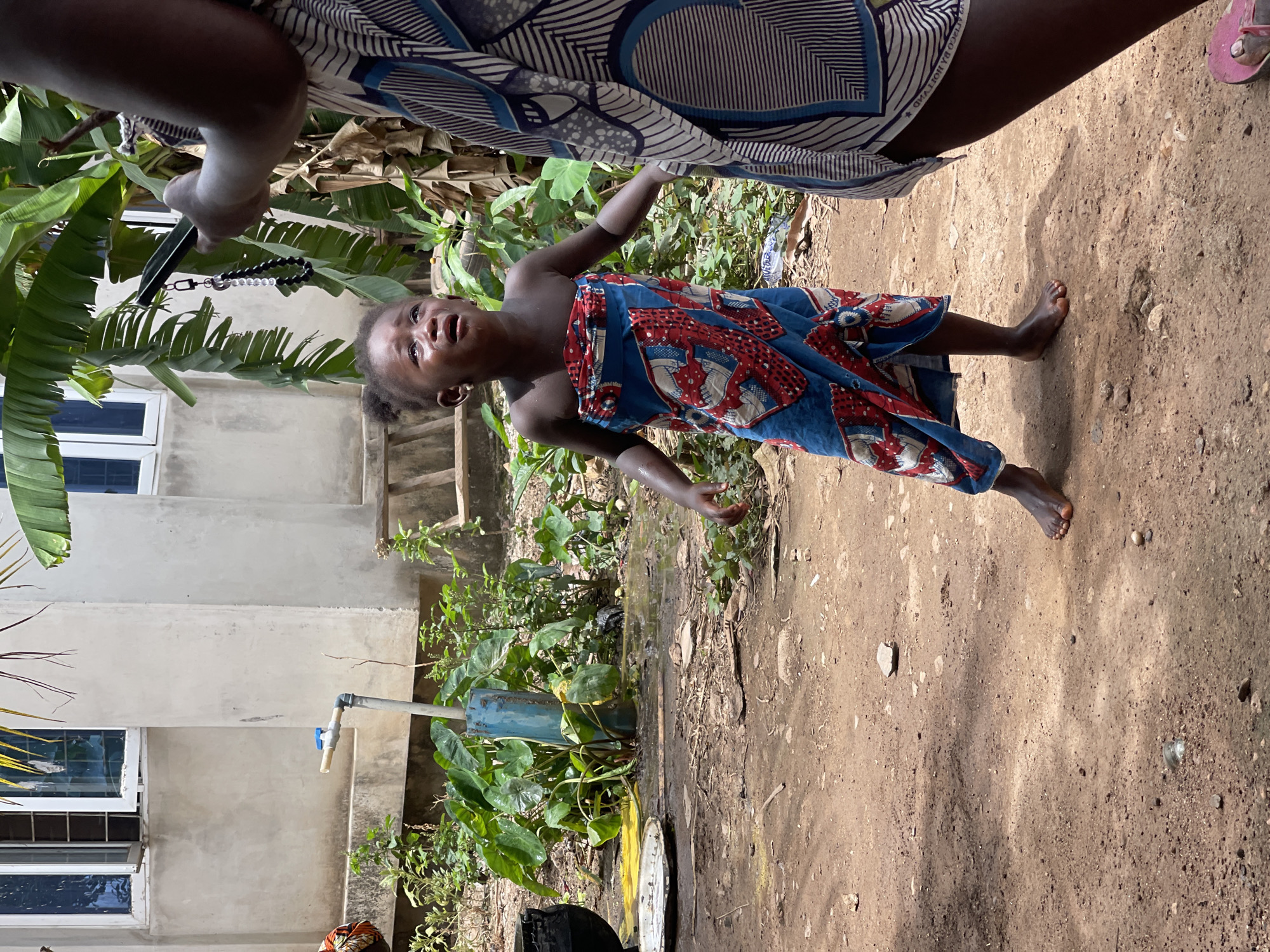 A young child wearing a colorful dress stands on a dirt surface near a building and a water pipe in an outdoor location. A person is holding a phone with a beaded strap. The surrounding environment includes vegetation and a structure with windows. The location appears to be in Akwanga, Nigeria.