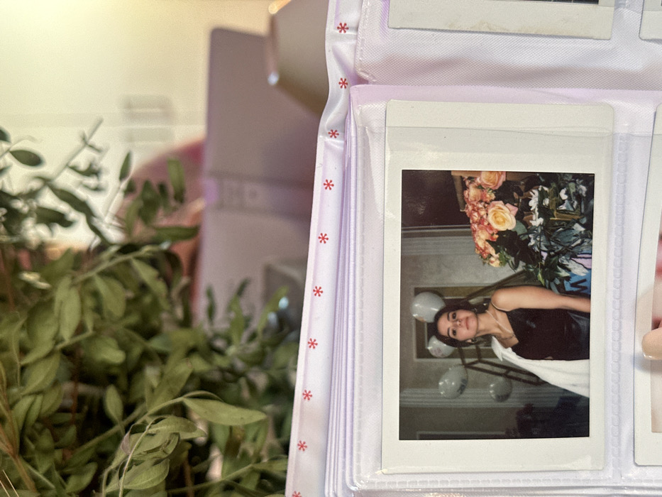 An instant print photograph, measuring approximately 7.5 cm by 10.5 cm, is displayed within a clear plastic sleeve of a light purple photo album. The album's upper edge features a white ribbon adorned with repeating red asterisk symbols. The photograph depicts a young female, approximately 20-30 years old, with dark hair and a neutral expression, looking directly at the viewer. She wears a black spaghetti-strap dress and has a white jacket draped over her left shoulder. A necklace with a pendant and earrings are visible. To the right of the subject is a large arrangement of light orange and pink roses with green foliage. In the background, to the left of the subject, several white balloons are visible. The setting within the photograph appears to be an indoor space with a dark wall. In the immediate foreground of the overall image, a dense cluster of green leaves and branches partially obstructs the upper portion of the photo album. The blurry background behind the album includes light-toned, indistinct objects, one of which appears to be a pink laptop or folder, and another, a light-colored lamp. The scene is located in 3-Y Uchastok, Russia.