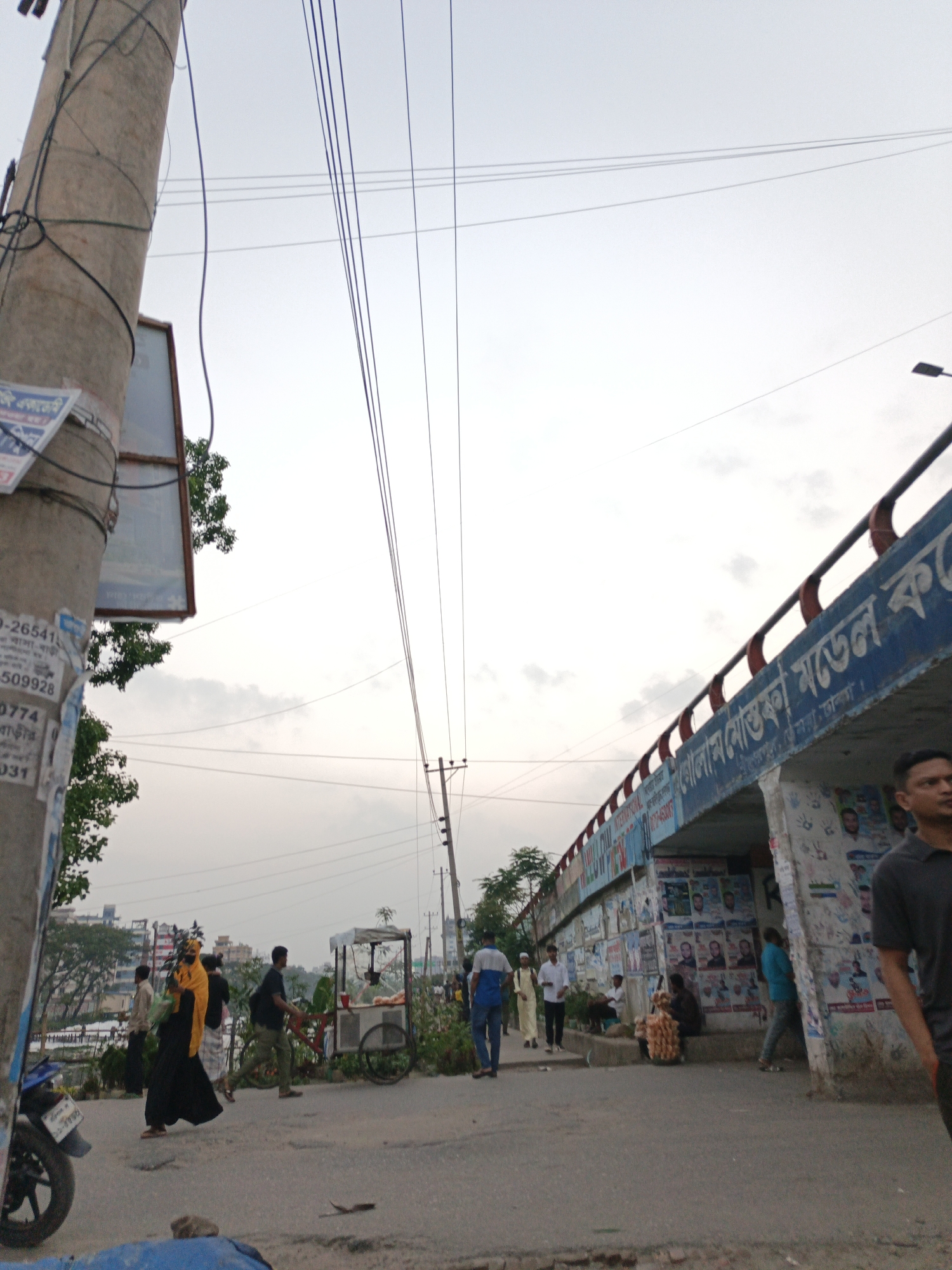 The image depicts a street scene in Dhaka, Bangladesh. A power pole with signage is on the left. Overhead, several power lines stretch across the sky. A bridge with graffiti and signage spans the street. People are walking and congregating near the bridge. A food vendor cart is also present. A motorcycle is parked on the left side of the street. The sky is overcast.