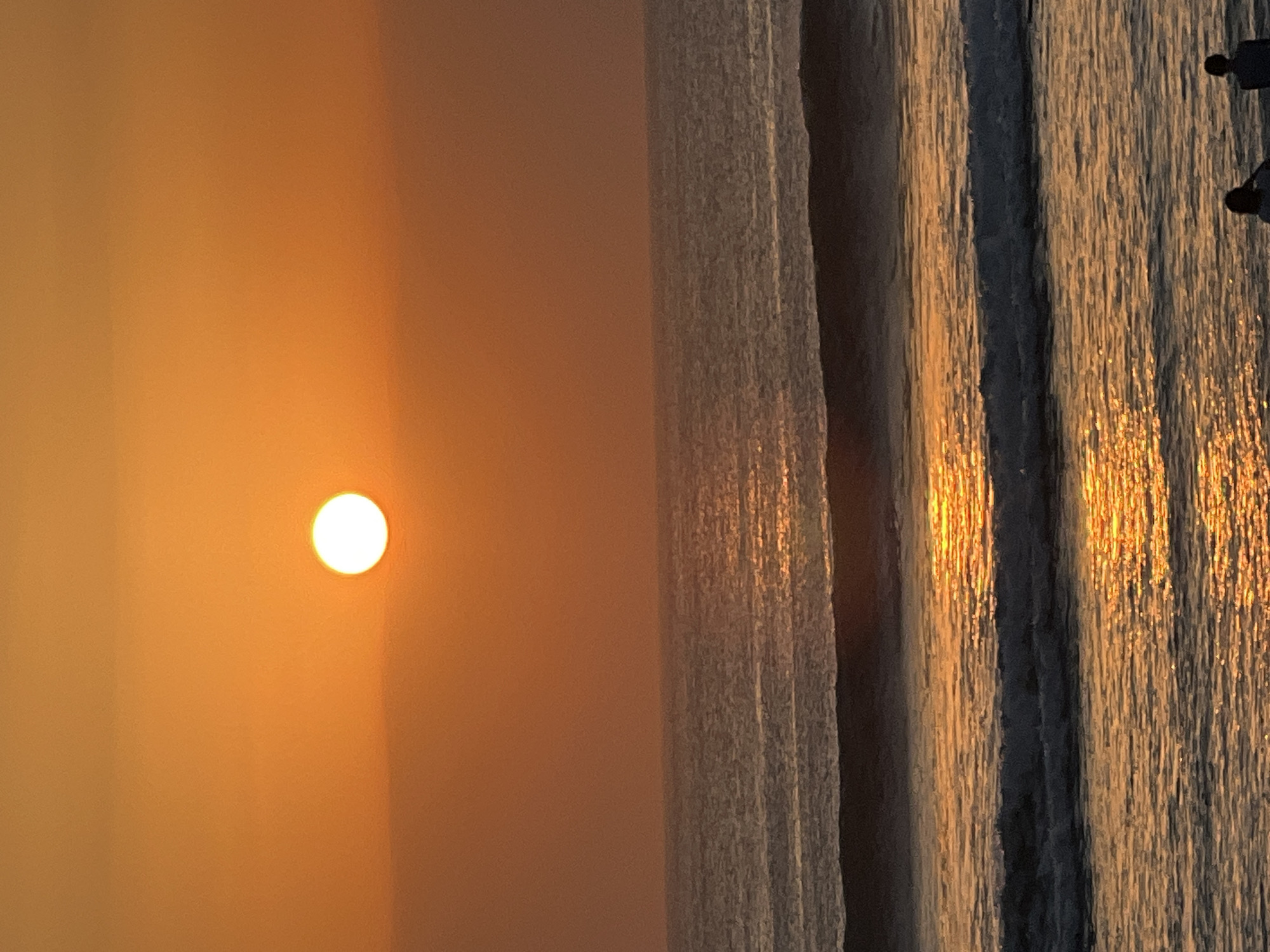 A wide-angle, vertical shot captures a sunset over the ocean in Kuta, Indonesia. The sun, a bright white orb, is positioned in the upper center of the frame, casting a warm orange glow across the sky and reflecting on the water. The ocean is calm with small waves breaking in the foreground, their crests highlighted by the setting sun. In the far foreground, two silhouetted figures are visible, facing the ocean. The overall scene is serene and peaceful, with the warm colors of the sunset dominating the composition.
