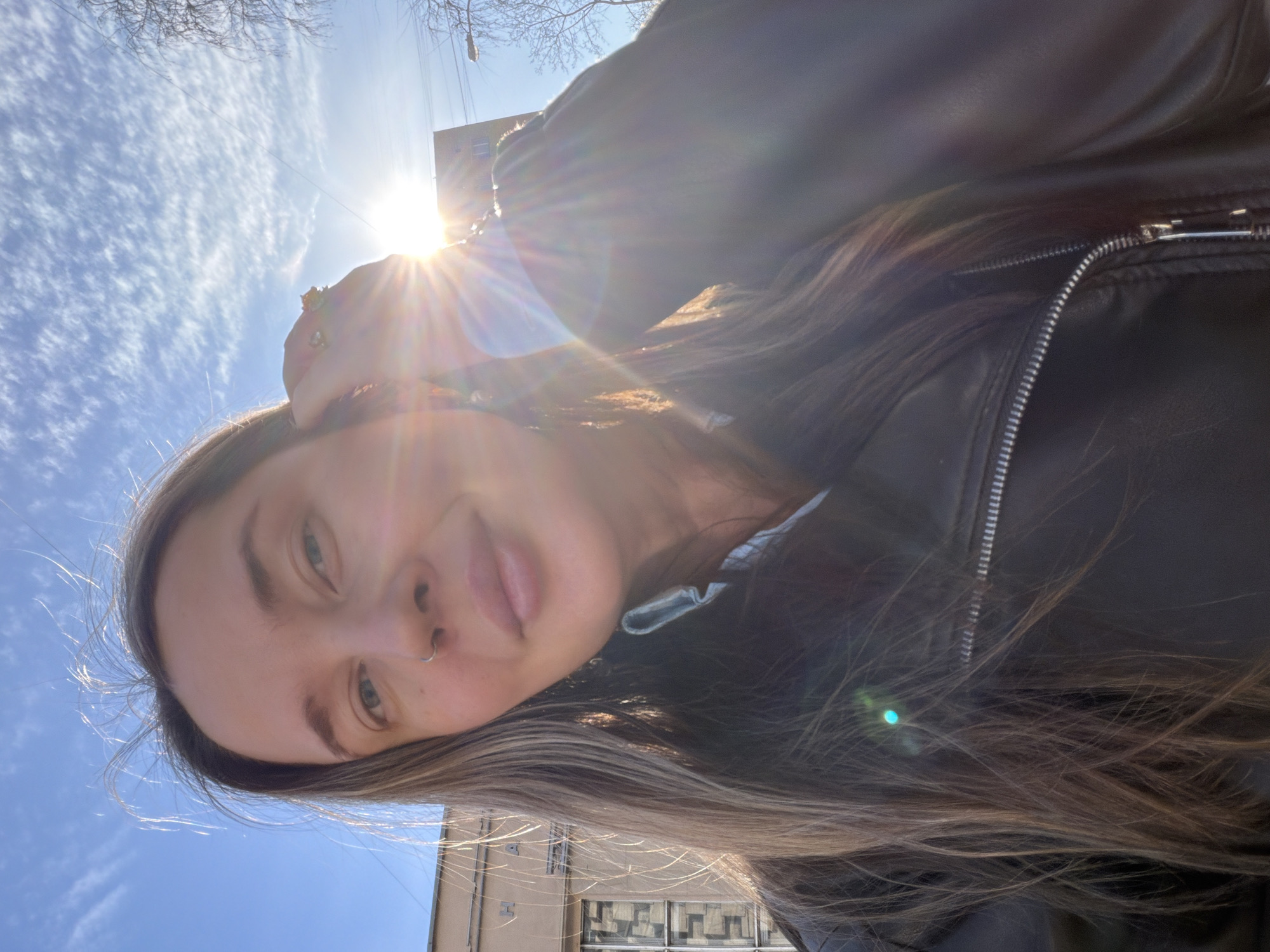 A selfie shot of a woman outdoors, likely in an urban setting. The woman has long brown hair and is wearing a black leather jacket. The sky is bright blue with some clouds, and the sun is shining brightly, causing lens flare in the image. In the background, a building is visible. The location is Moscow, Russia.