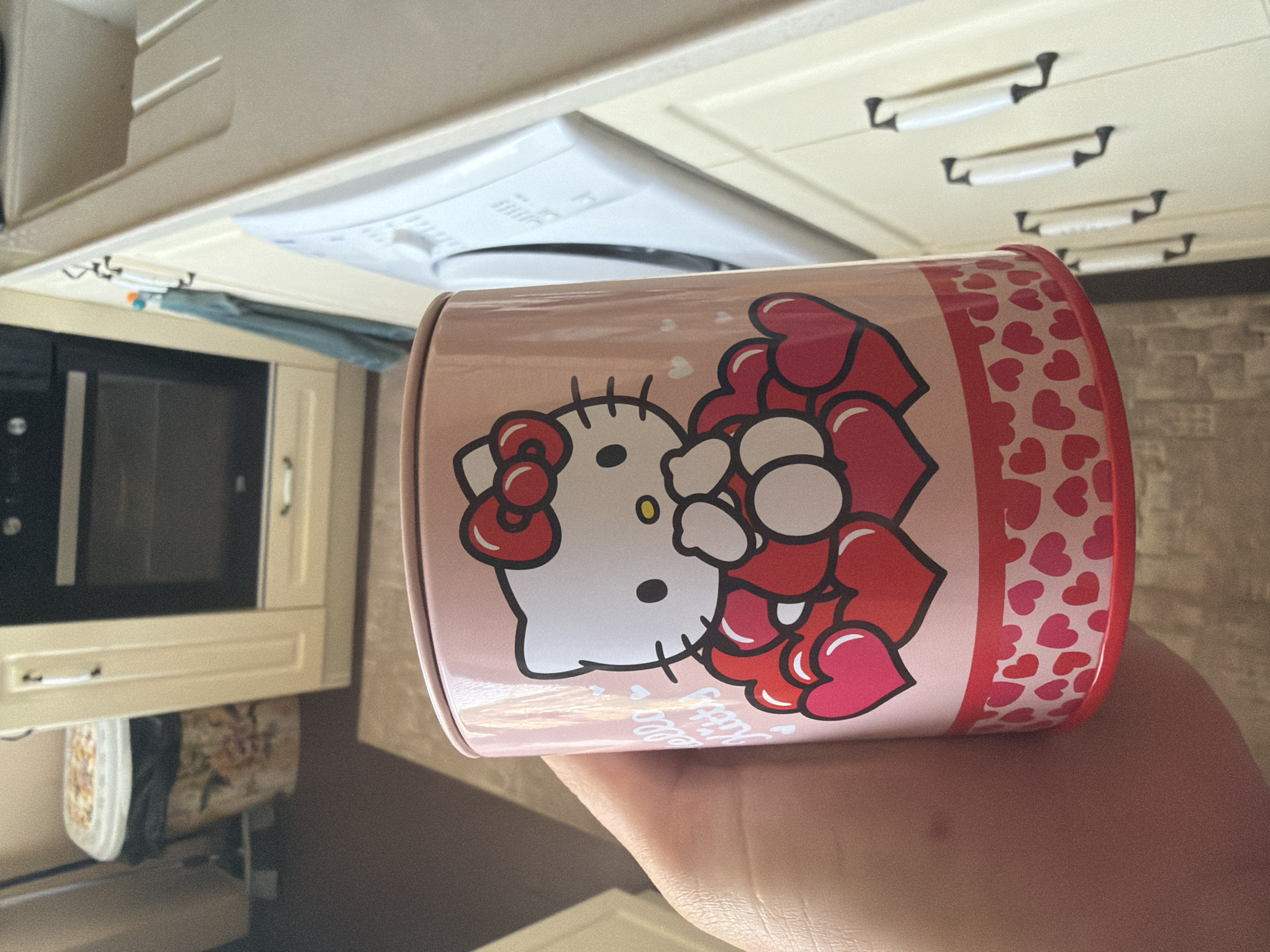 The image, captured in a sideways orientation, depicts a person's hand holding a pink cylindrical Hello Kitty container within a domestic kitchen setting.
The container is the central focus, featuring a large graphic of Hello Kitty's face with her characteristic red bow and yellow nose. She is surrounded by numerous red and pink hearts of various sizes, with a vertical band of smaller hearts running along the side of the container. Partially visible white text below Hello Kitty's face reads "Hello Kitty". The container appears to be made of a reflective material like tin.
The background reveals a well-lit kitchen. To the left, a built-in microwave oven with a dark door is integrated into light-colored cabinetry. Below the microwave, a patterned fabric item, possibly a dish towel or bag with a floral design, is visible. Above the microwave, a light-colored cloth hangs from hooks attached to the underside of the upper cabinets. These upper cabinets are also light-colored, featuring dark, possibly black, handles on multiple drawers or doors. On the upper right, a white kitchen appliance, likely a range hood or part of a cooking unit, is visible with control knobs. The wall in the background has a warm, neutral tone and a textured appearance, possibly resembling brick or stone.
The activity shown is simply someone holding the Hello Kitty container. The scene suggests a casual moment indoors, likely during the daytime or in a brightly lit kitchen in Shumen, Bulgaria.