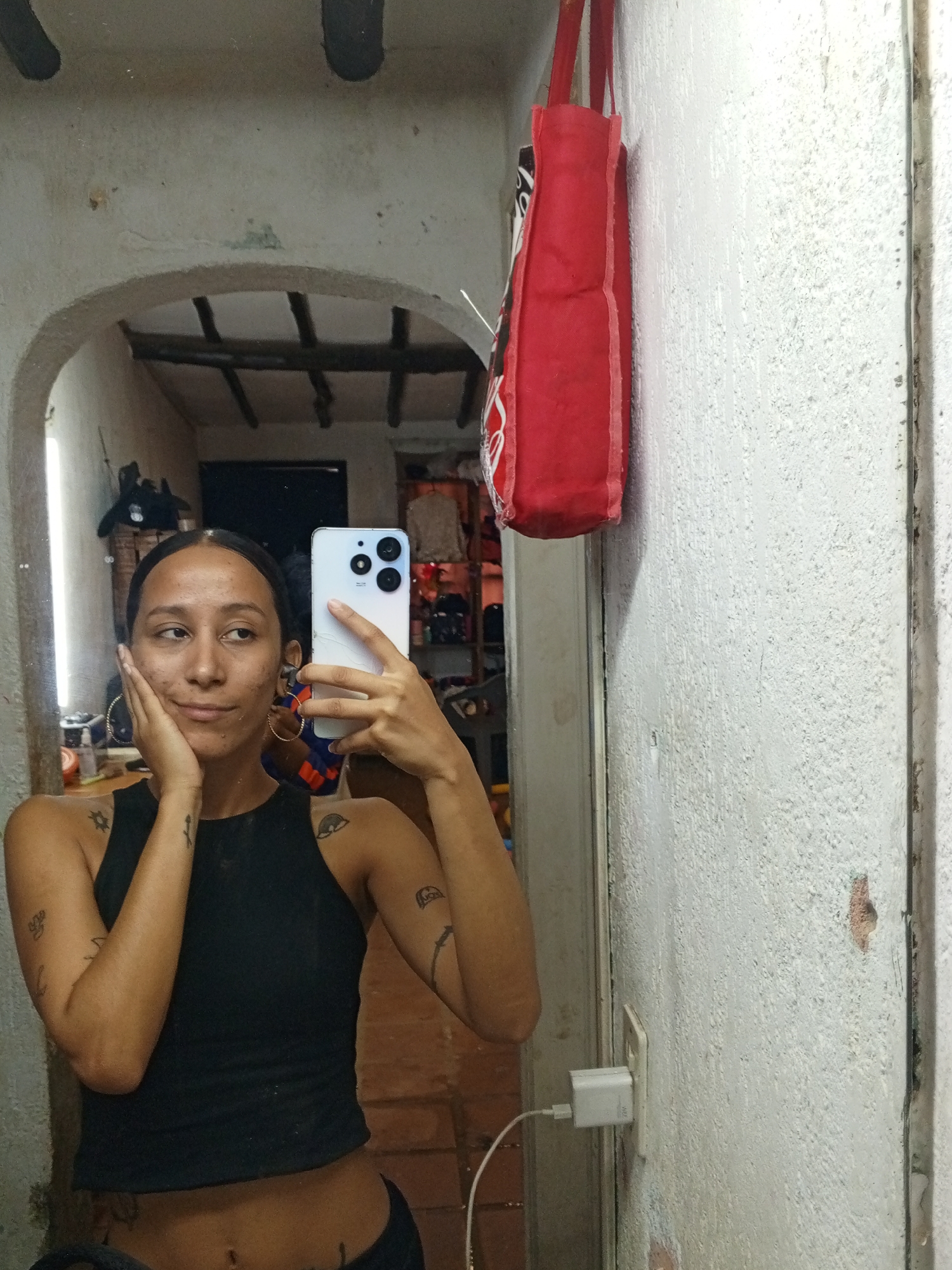 A young adult female is taking a selfie in a mirror. She has dark hair pulled back, and wears a black tank top. Tattoos are visible on her arms and abdomen. She is holding a white smartphone with multiple camera lenses. Her left hand is touching her cheek.
The background shows a room interior. The mirror is framed by an archway with exposed beams. There is a red tote bag hanging on the right side of the mirror. A wall with a power outlet and charger is also visible on the right side of the frame. The location appears to be a domestic interior in Porlamar, Venezuela.