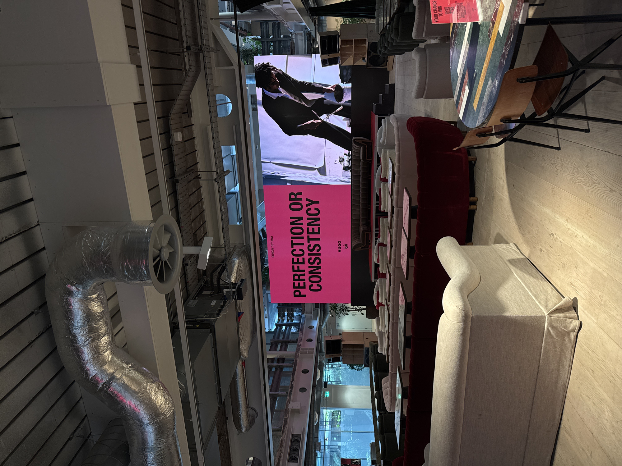 The interior of a space in London, United Kingdom, is depicted. A screen is displaying a man in a dark suit. In front of the screen hangs a pink sign with white text. The words on the sign read "PERFECTION OR CONSISTENCY". Rows of seating and tables, which appear to be arranged for an event or presentation, fill the space. A large, industrial-looking duct with a fan is mounted to the ceiling. The floor is a light wooden laminate. The lighting appears to be a mix of natural light from the windows and artificial light from the ceiling.