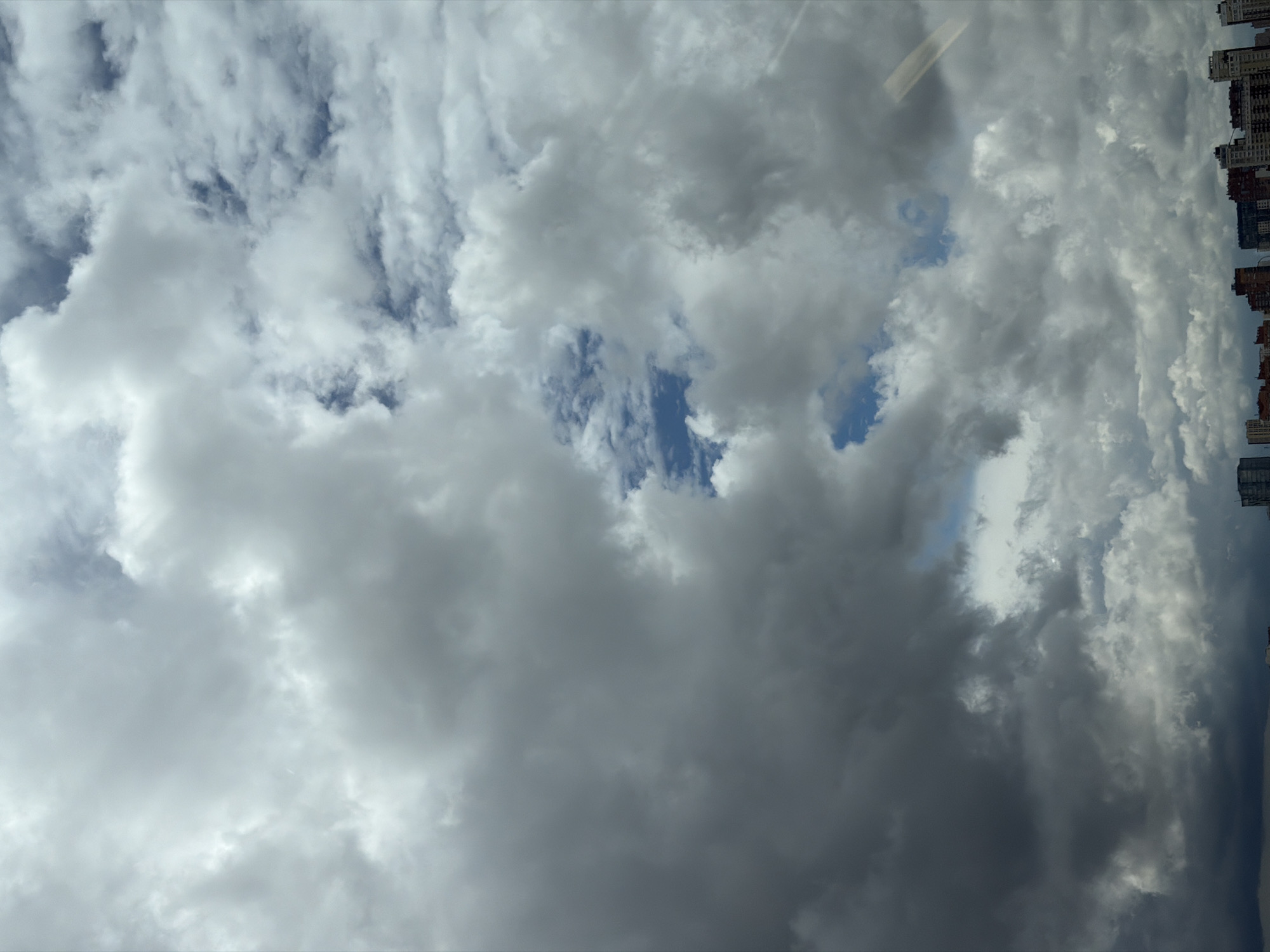 The image depicts a cloudy sky over a city. The clouds are varied in appearance, with some being dark and heavy, suggesting an approaching storm, while others are lighter and more scattered, allowing glimpses of blue sky. The lower portion of the image shows a cityscape with buildings of varying heights and architectural styles. The buildings are dense and appear to be residential or mixed-use. The overall impression is of an urban environment under a dynamic sky. The specific location within Moscow, Russia, cannot be determined from the visual information alone, as the image lacks any identifying landmarks or distinctive features that would pinpoint a particular district or area. The scene is static, with no visible people or ongoing actions.