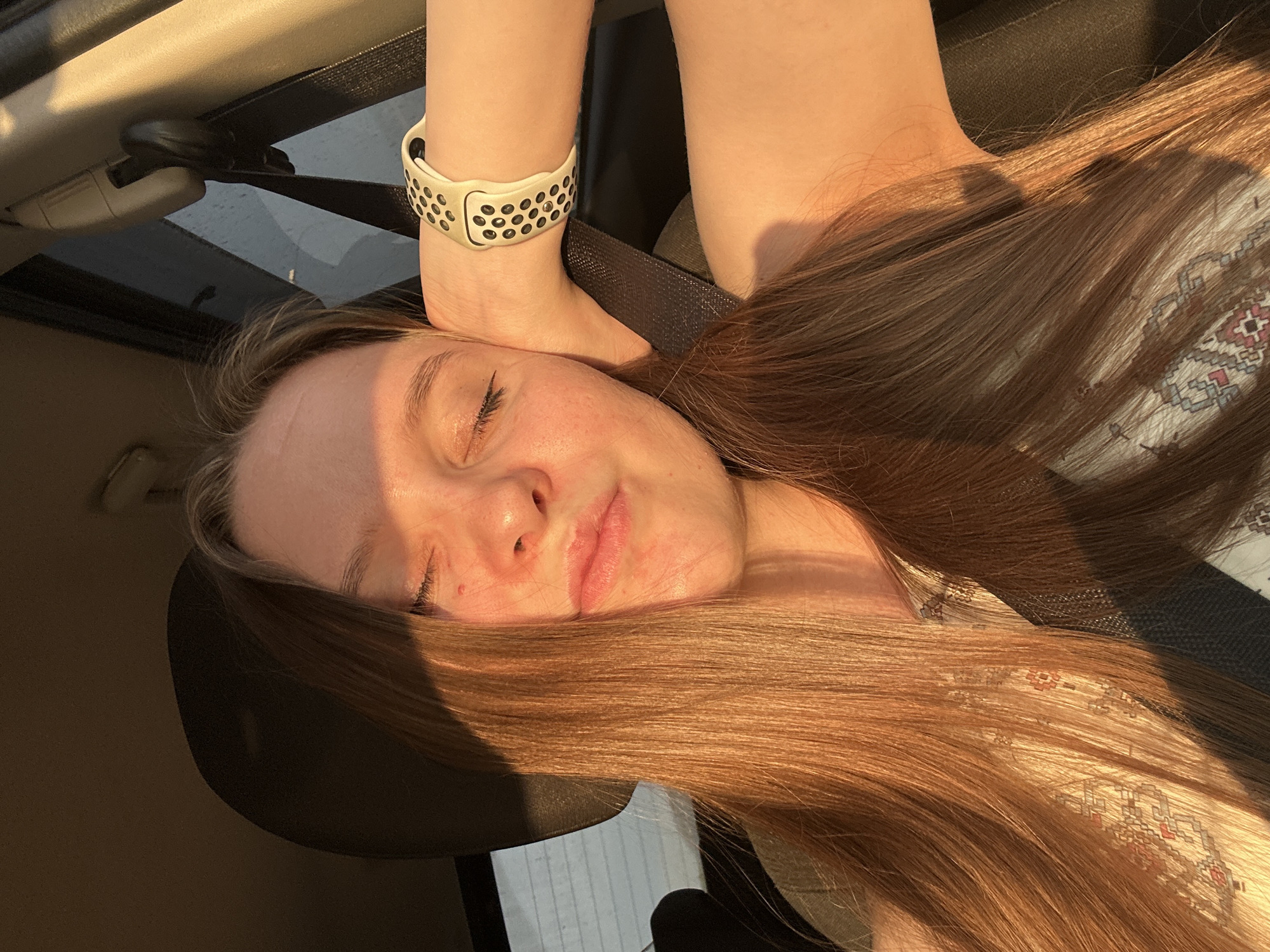 A young woman with long, reddish-brown hair is captured in a close-up selfie taken inside a vehicle. She is leaning her head on her hand, with her eyes closed and her lips slightly parted. The setting sun casts a warm, golden light across her face and hair. She is wearing a patterned top and a smartwatch on her left wrist. The background shows a seatbelt and the interior of a car, with a window visible behind her. The image evokes a sense of peace and relaxation. The image does not provide any specific indicators to determine its location within the city of Chaikovsky, Russia.