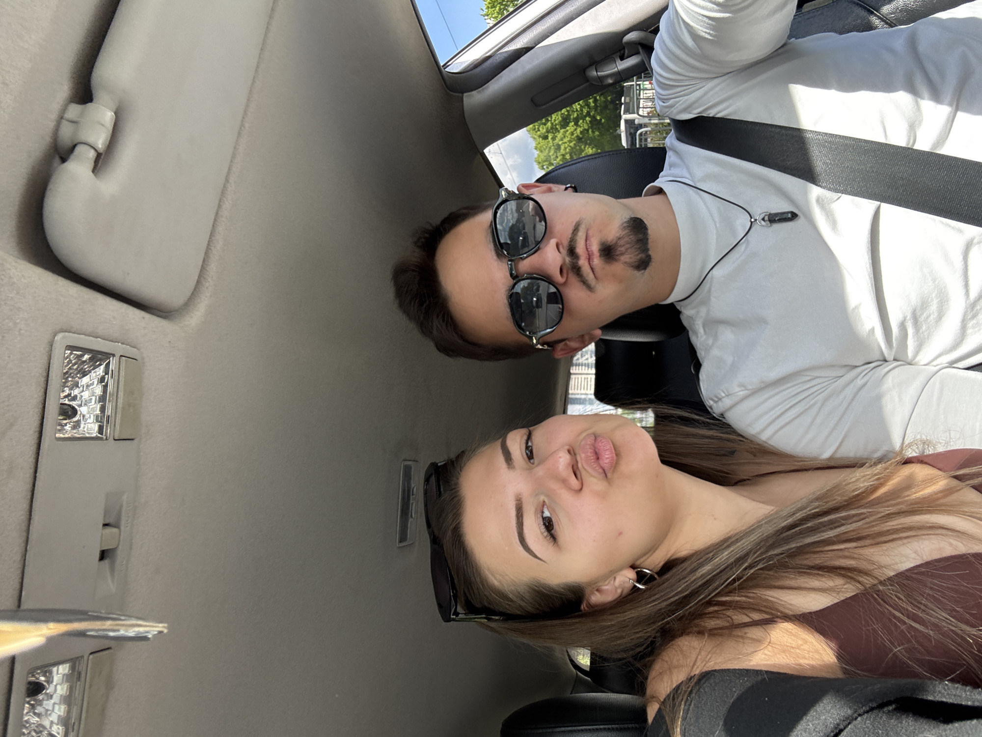 The image depicts two individuals in a car. The man, positioned in the upper portion of the frame, wears sunglasses and a white long-sleeved shirt with a black seatbelt. He has dark hair and a facial hair style that includes a mustache and goatee. He is wearing a necklace. The woman is in the lower portion of the frame, she has long brown hair and wears a brown top. She is making a kissing face. It is daytime and the lighting indicates the presence of natural light. The car's interior is light gray, and visible elements include the sun visor, interior lights, and ceiling. The precise location in Sofia, Bulgaria, is not identifiable from this image alone.